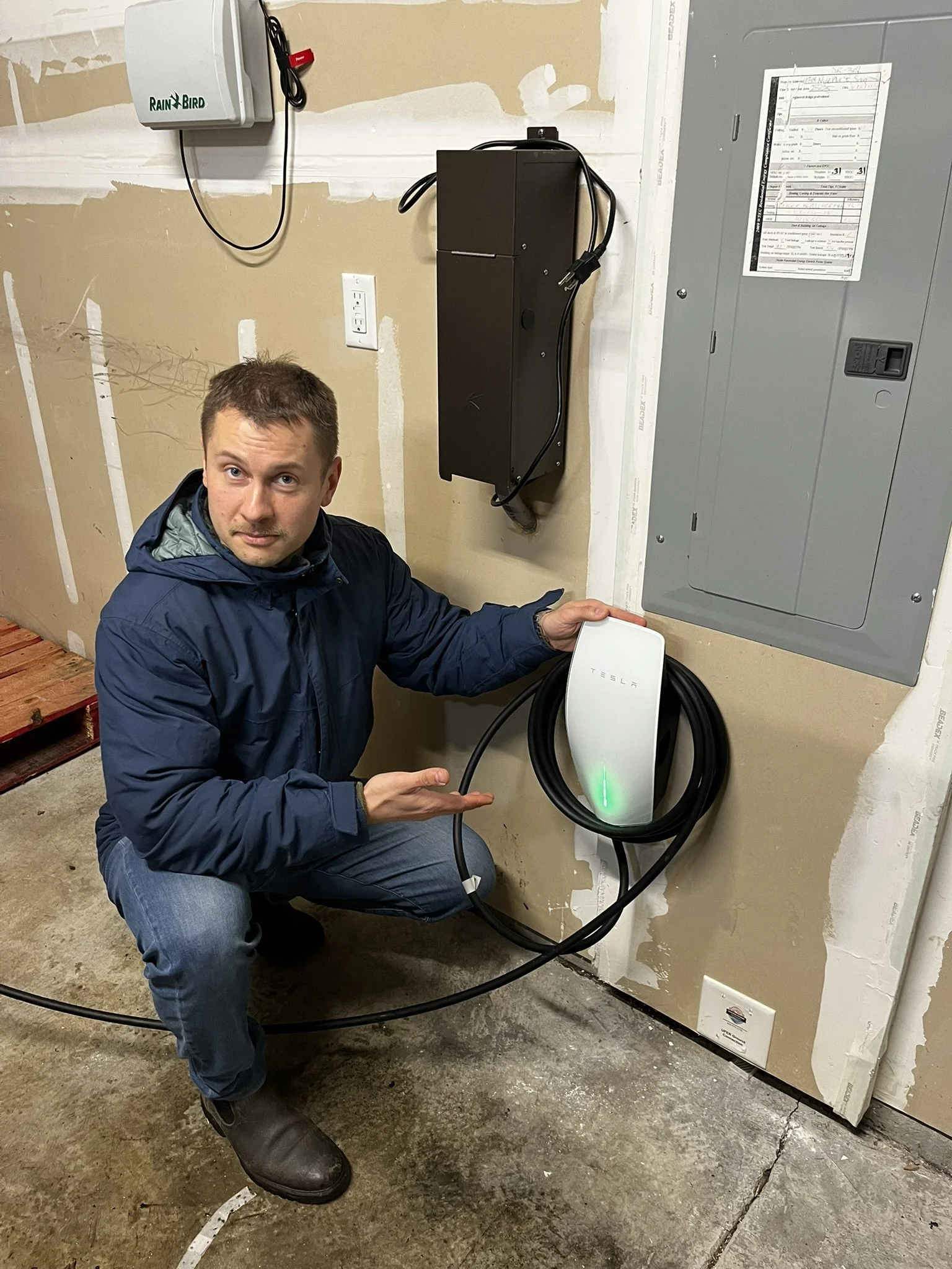 Man crouching next to an electric vehicle charging station, holding the charging unit, in a garage with unfinished walls and electrical panels.