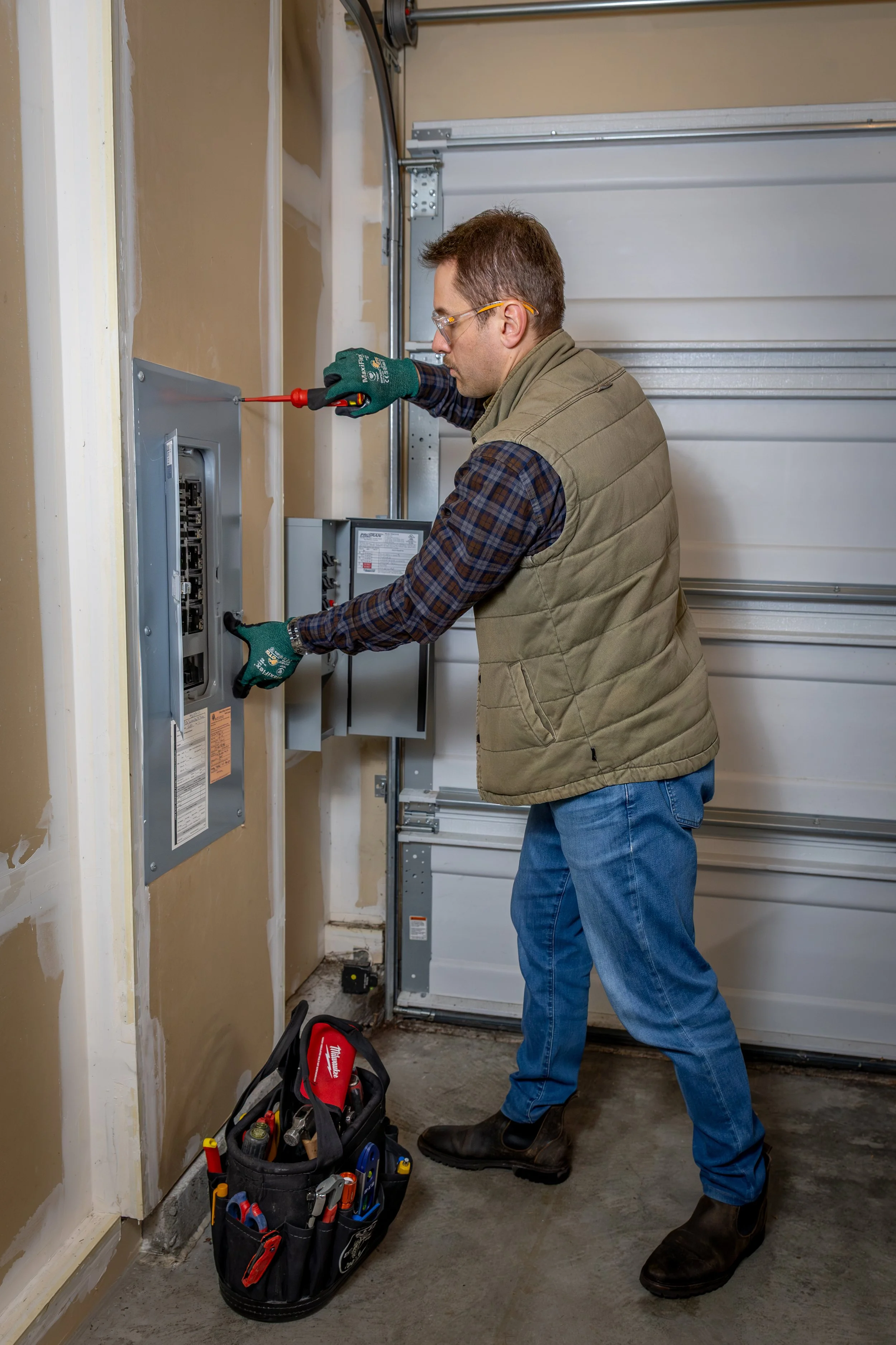 A man wearing safety glasses, gloves, a green quilted vest, and jeans is working on an electrical panel in a garage. He is using a screwdriver and has a tool bag with various tools on the floor nearby.