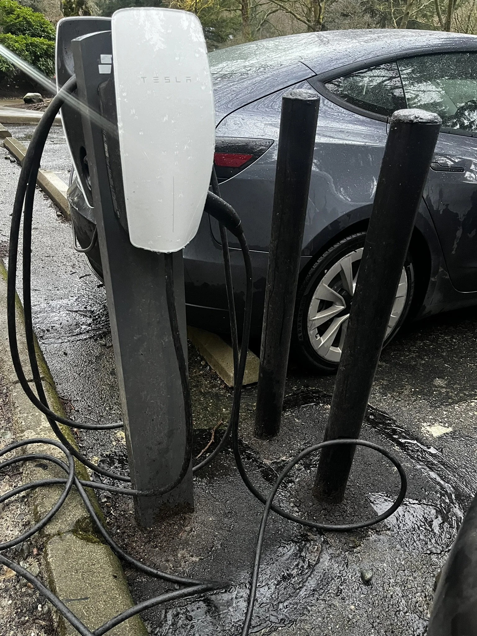 Electric vehicle charging station next to a gray Tesla parked in a wet parking lot.