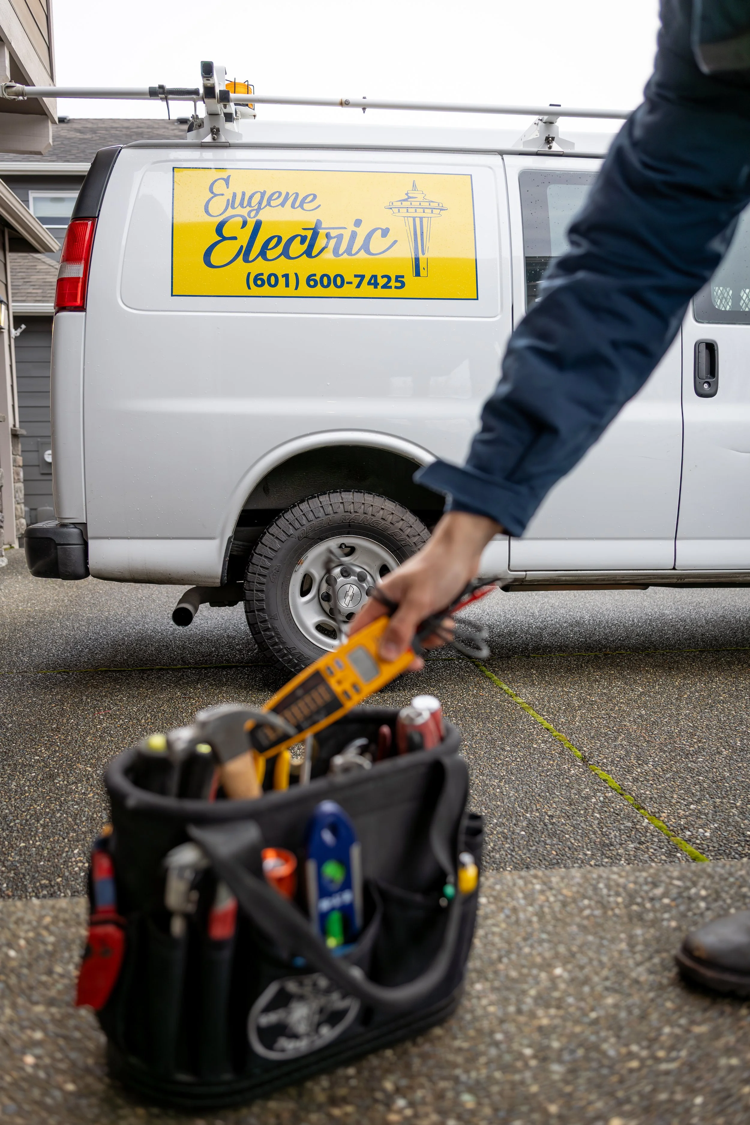 A technician using a multimeter on a vehicle with a sign that reads 'Eugene Electric' and a phone number, with a tool bag in the foreground.