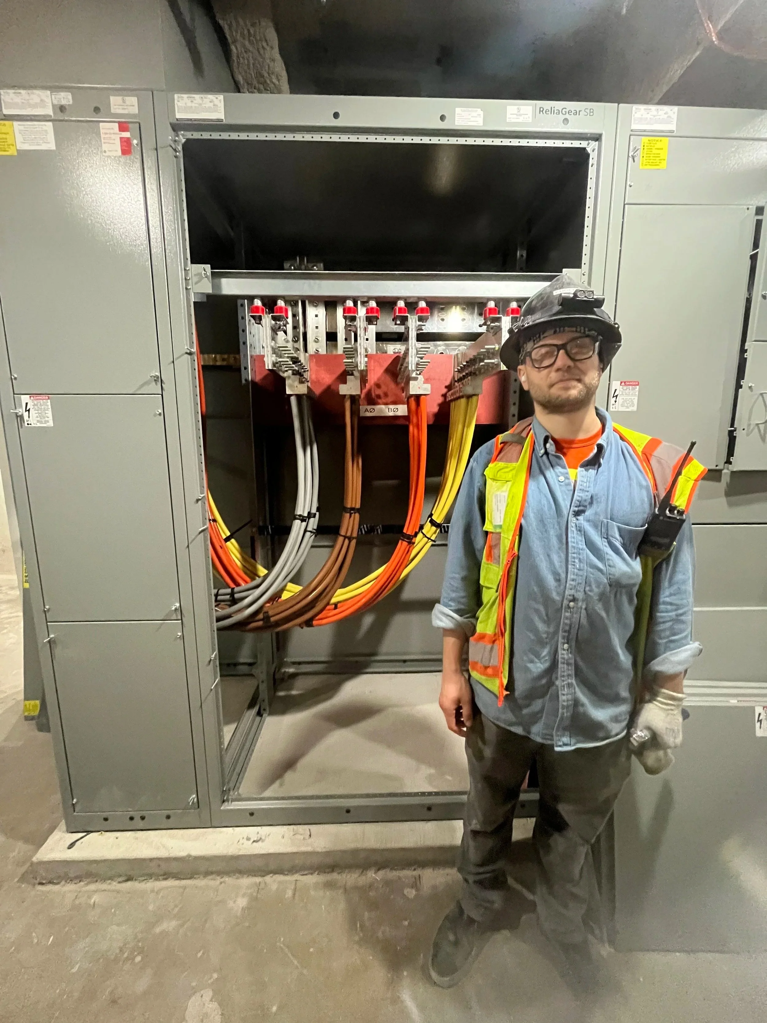 A worker standing in front of an electrical substation with multicolored cables and switches, wearing safety gear including a helmet and high-visibility vest.