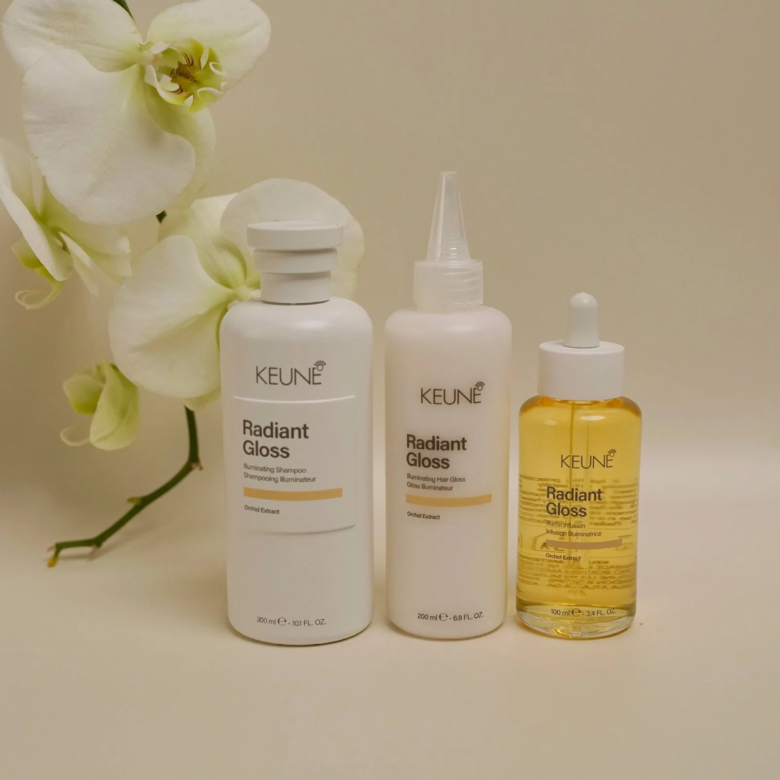 Three beauty products labeled KEUNE Radiant Gloss, with white and yellow packaging, placed in front of a white orchid flower on a plain background.