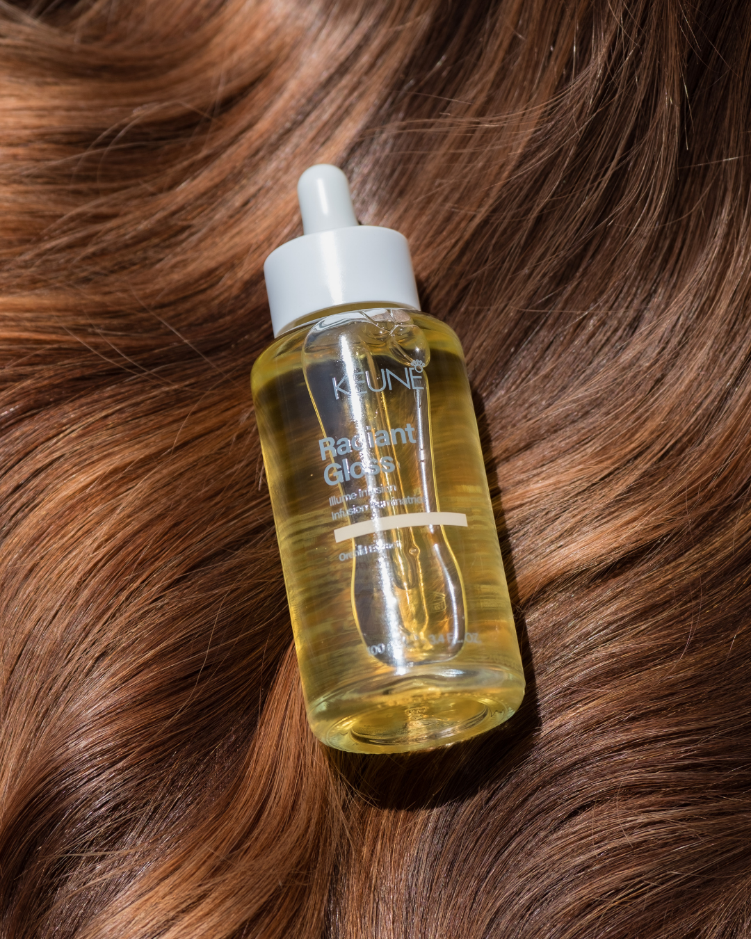 A bottle of Keune Radiant Gloss hair treatment on wavy, reddish-brown hair.
