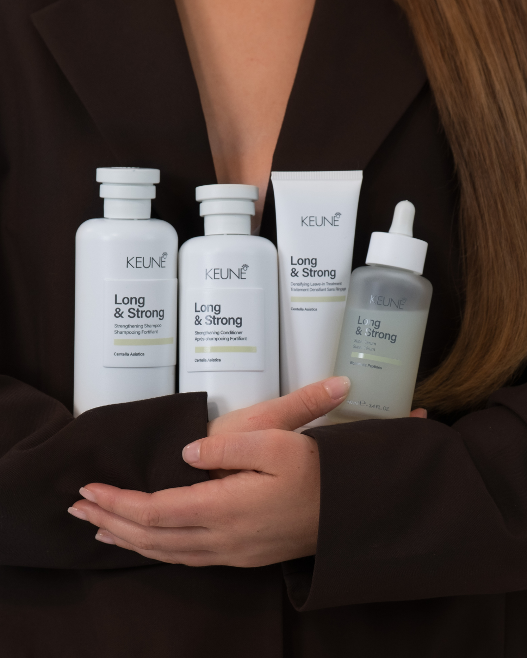 A person holding four bottles of KEUNE Long & Strong hair care products, including shampoo, conditioner, and treatment, against a dark background.