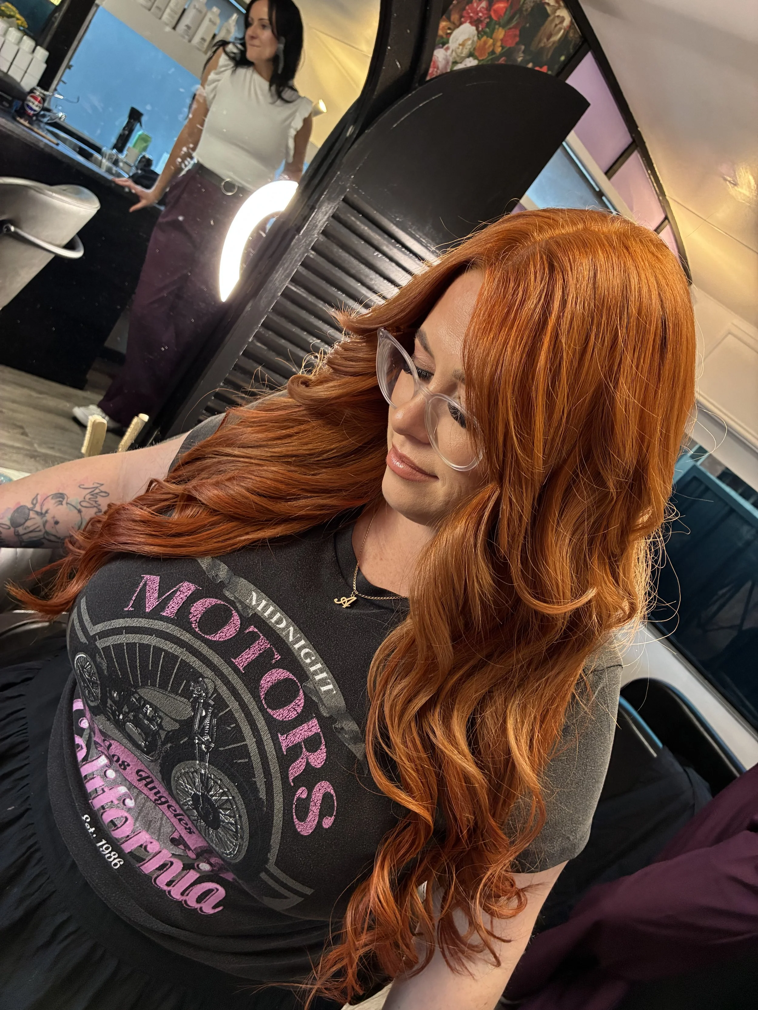 A woman with long, wavy red hair, glasses, and a tattoo on her arm sitting indoors. Hair extensions rolleston