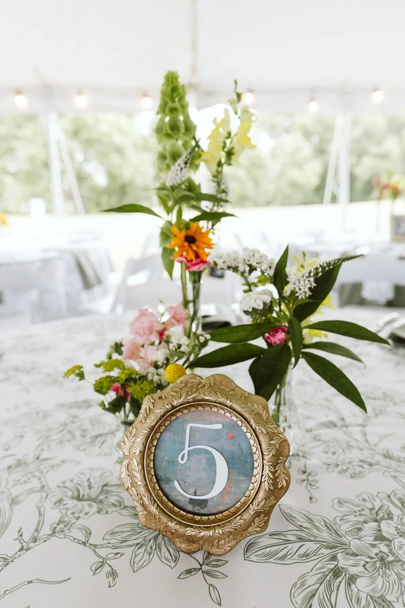 A decorative table number in a gold ornate frame with the number 5, placed on a floral tablecloth, with a centerpiece of mixed fresh flowers including pink, white, yellow, and green blooms in small vases in the background.
