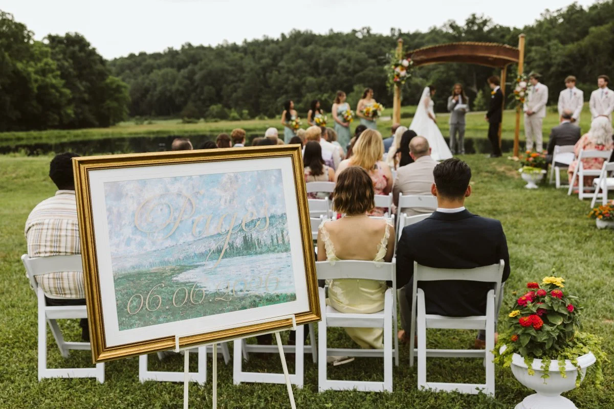 An outdoor wedding ceremony taking place by a river, with the bride and groom exchanging vows under a wooden arch, guests seated on white chairs, and a framed sign with the word 'Peace' and a date in the foreground.