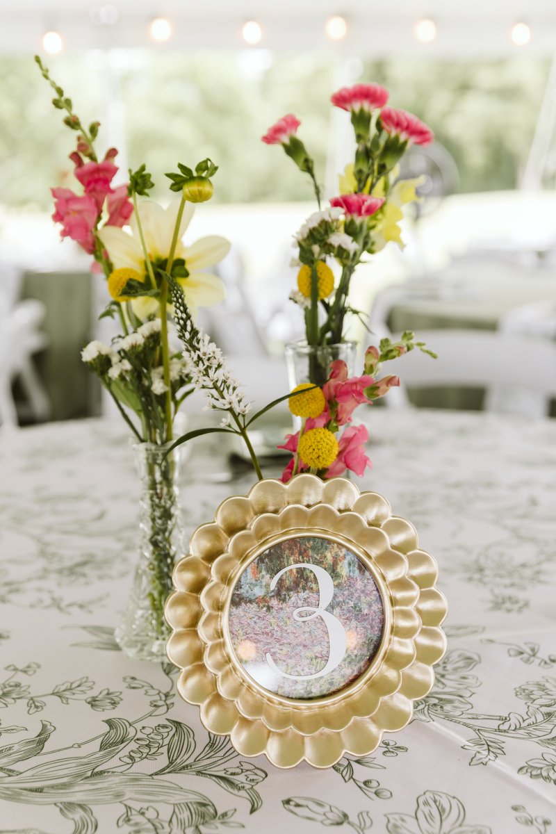 Table centerpiece with colorful wildflowers in glass vases, and a gold scalloped table number frame with the number 3, set on a white tablecloth with a floral pattern.