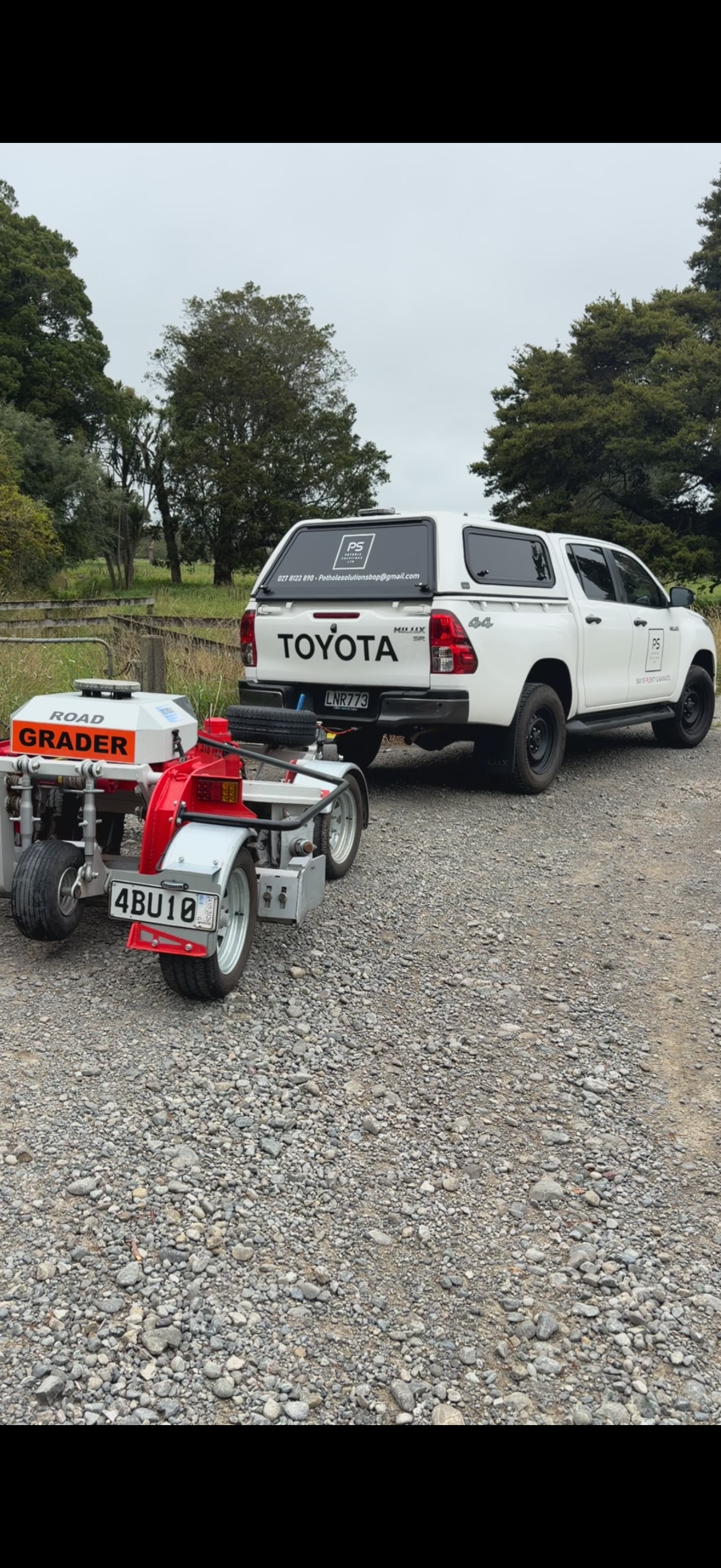 A white Toyota pickup truck with a canopy parked on a gravel road, with a small white and red grader vehicle attached in front of it.