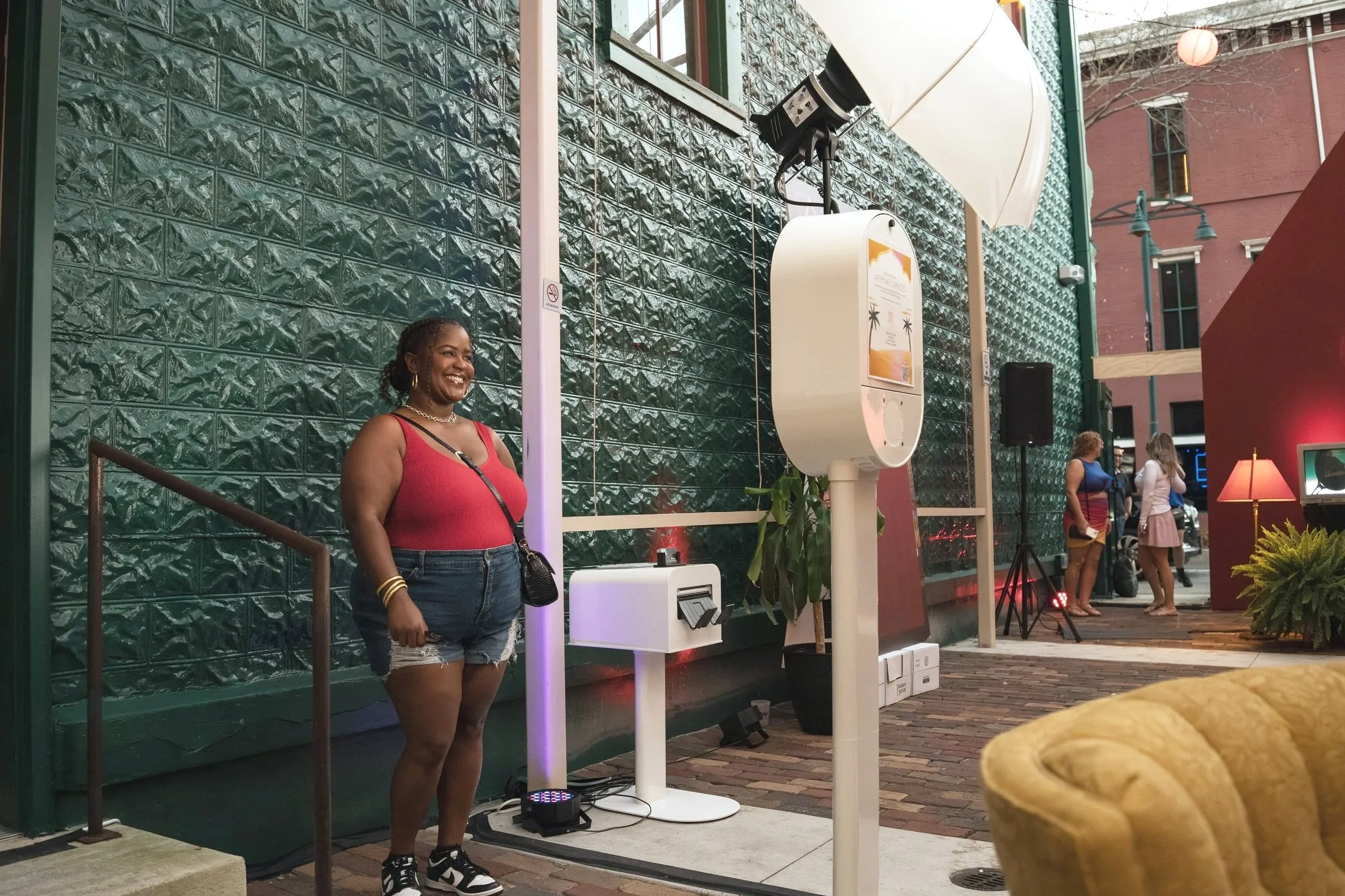 A woman in a red tank top and denim shorts is smiling while posing outside a building with a green textured wall. There is a photo booth with lighting and a digital screen nearby. In the background, three women are standing and talking.