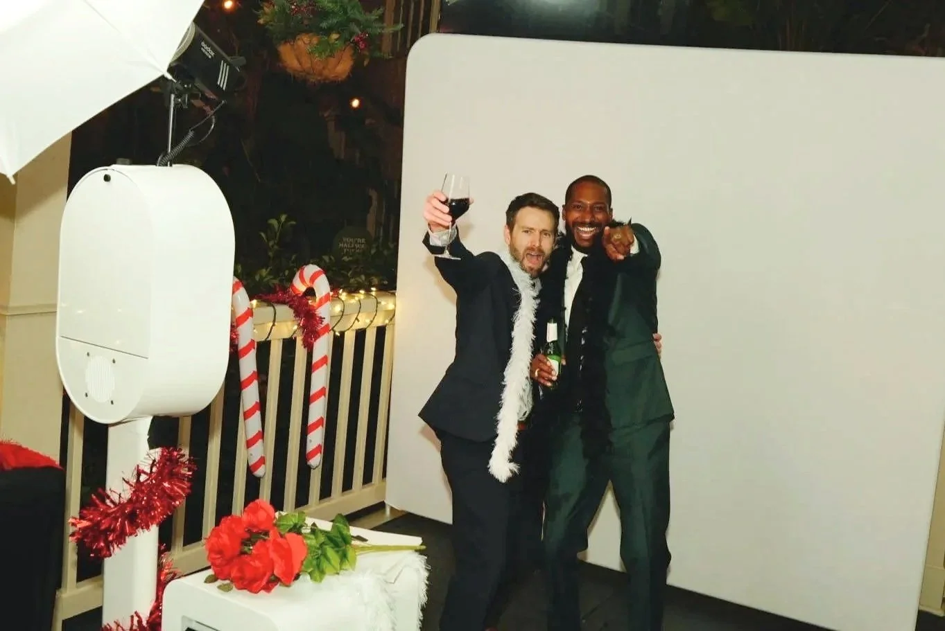 Two men dressed in tuxedos celebrating with drinks at a holiday party, standing in front of a white backdrop, decorated with Christmas ornaments, candy canes, and flowers.