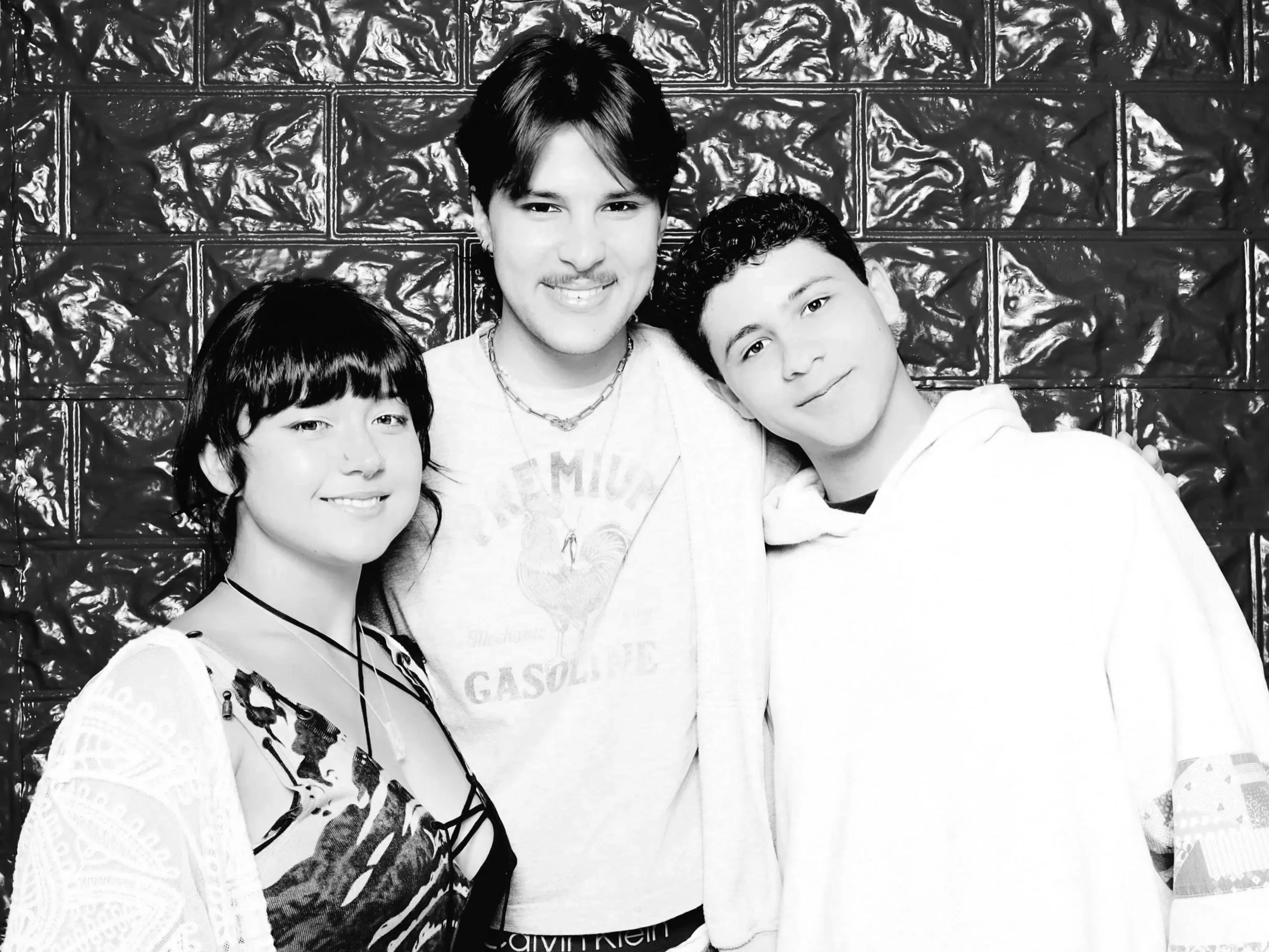 Black and white photo of three smiling teenagers standing close together in front of a textured wall, with the teen on the right slightly leaning on the middle teen.