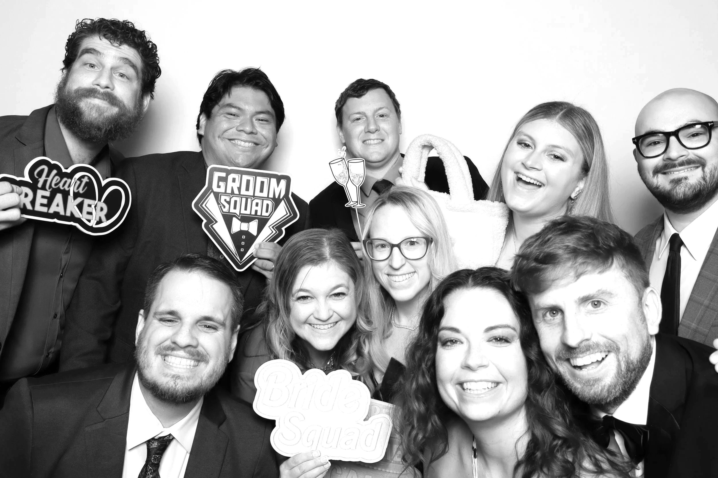 Group of people at a wedding celebration posing with playful signs and props, including a heartbreaker sign, groomsquad sign, bride squad sign, and drawing with champagne glasses. They are smiling and dressed in formal attire.