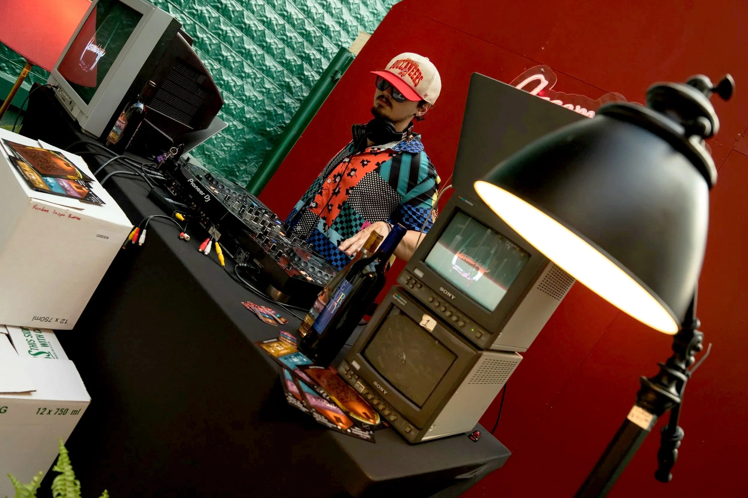 A DJ wearing sunglasses, a colorful patterned shirt, and a baseball cap is standing behind a DJ setup with turntables, a mixer, and computer equipment. The setup is on a black table with books, brochures, and a couple of bottles nearby. The backgroun