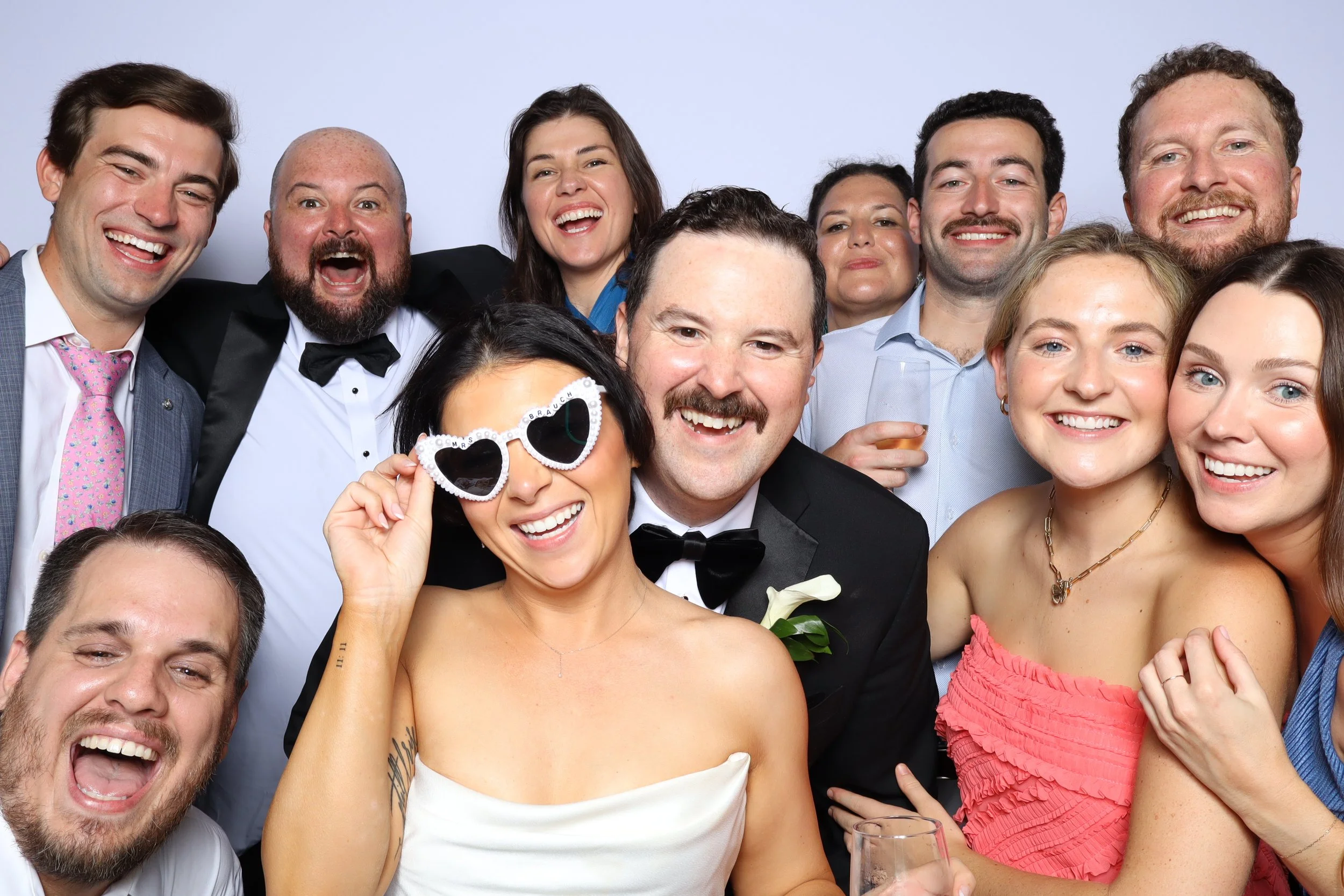 Group of ten friends at a celebration, smiling, dressed in formal and semi-formal attire, some holding glasses, with one person wearing heart-shaped sunglasses.