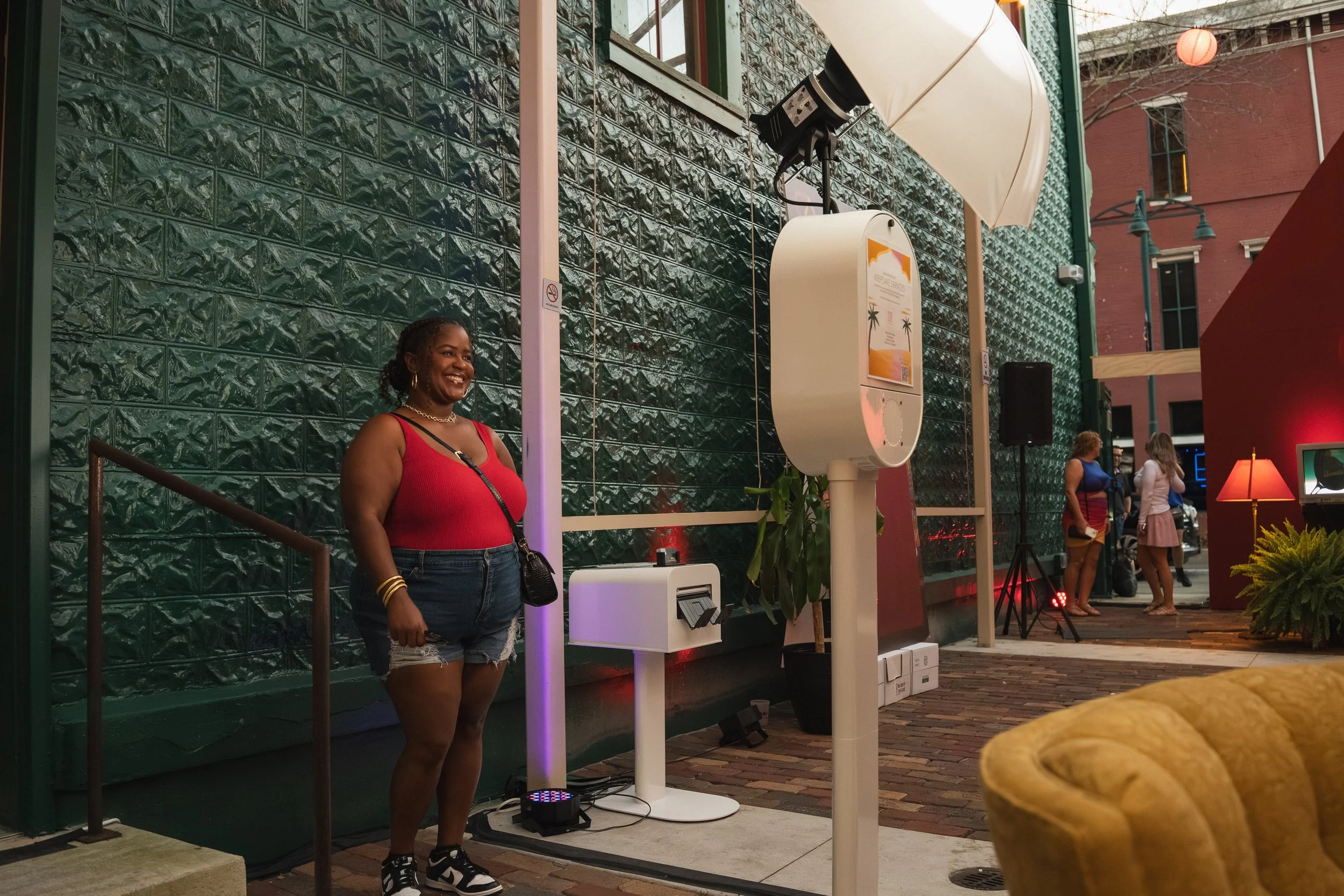 A woman in a red tank top and denim shorts smiling at an outdoor event, with a green textured wall behind her and people in the background