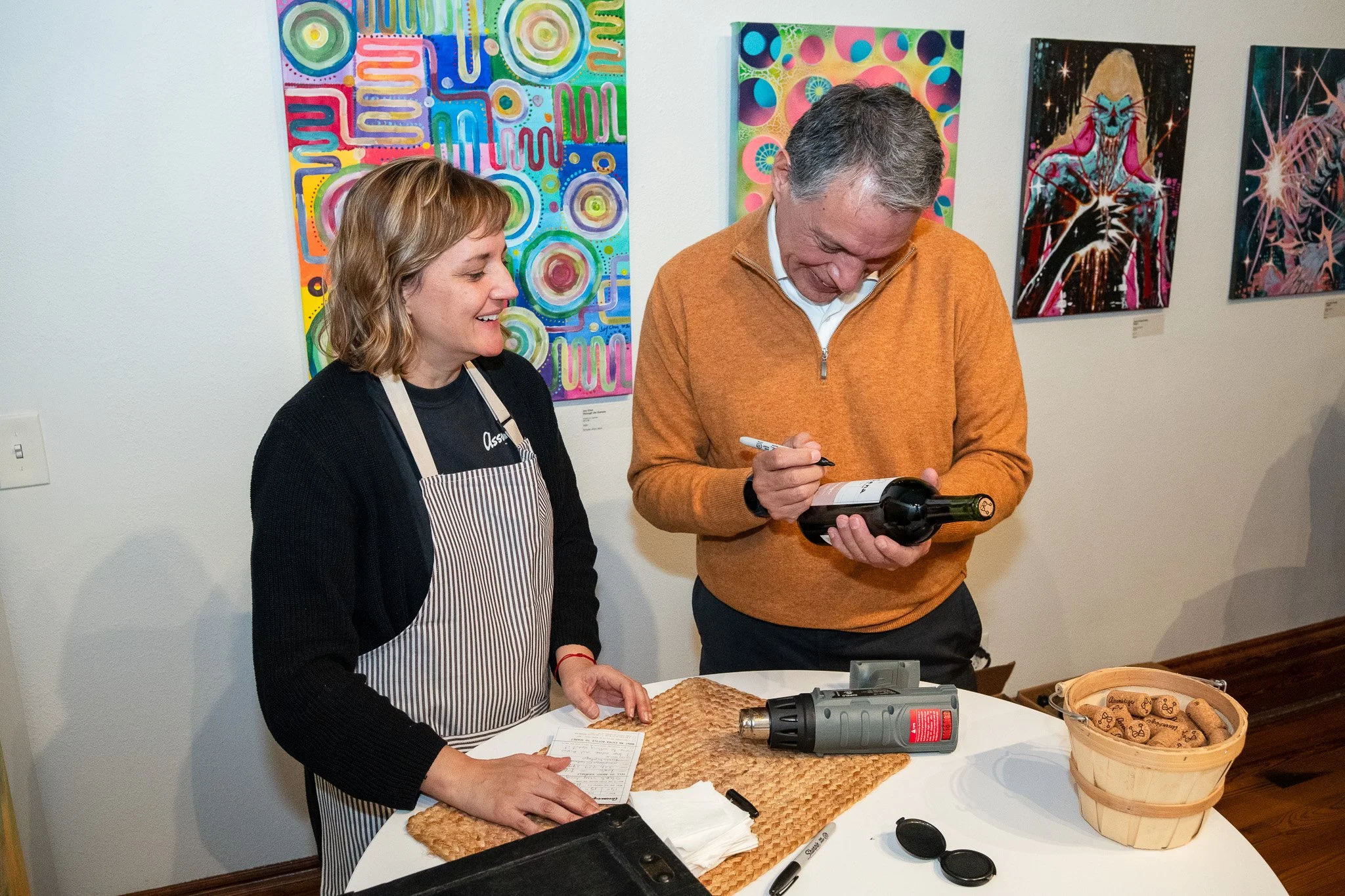 A woman and a man are standing at a table in an art gallery. The woman, wearing a black sweater and apron, is smiling and looking at the man. The man, wearing an orange sweater, is signing a wine bottle he is holding. On the table, there are corks, a