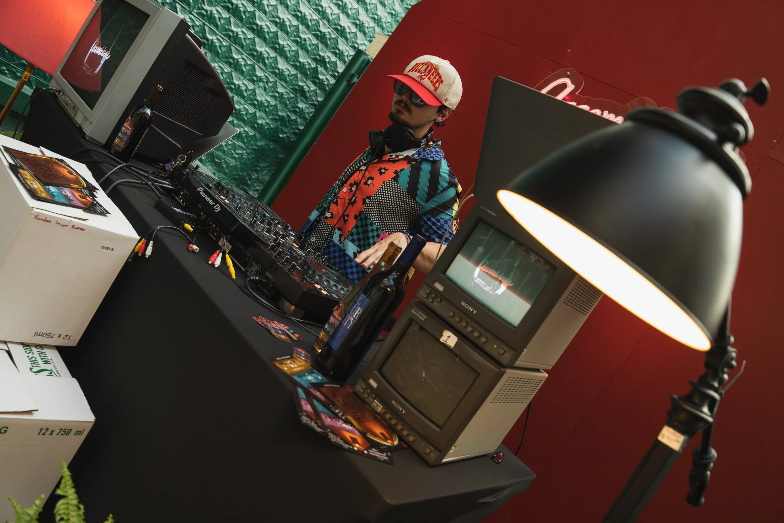 A DJ wearing sunglasses, a colorful patterned shirt, and a baseball cap is standing behind a DJ setup with turntables, a mixer, and computer equipment. The setup is on a black table with books, brochures, and a couple of bottles nearby. The backgroun