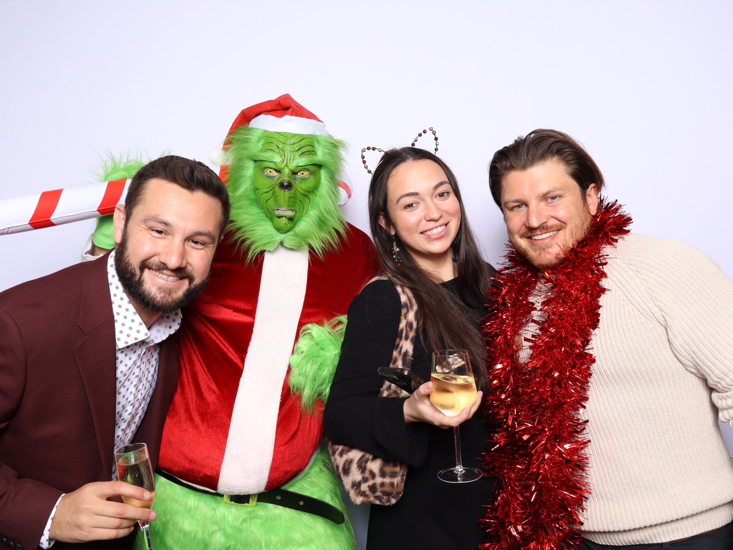 Four people at a holiday party, one dressed as the Grinch with a green face, red Santa hat, and red coat. The other three are smiling, holding drinks, with festive accessories like a red tinsel garland and animal ears headband.