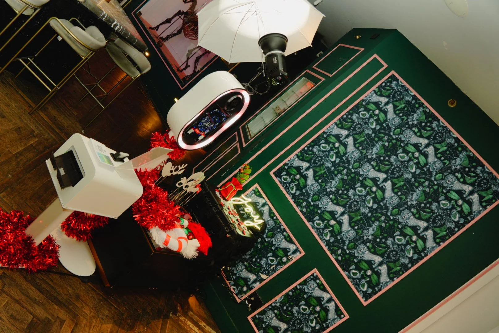 Holiday-themed indoor space with a white camera and a smartphone on a black surface, decorated with red Christmas garland and ornaments, against a green wall with patterned panels, and a neon 'Love' sign, illuminated by studio lighting.