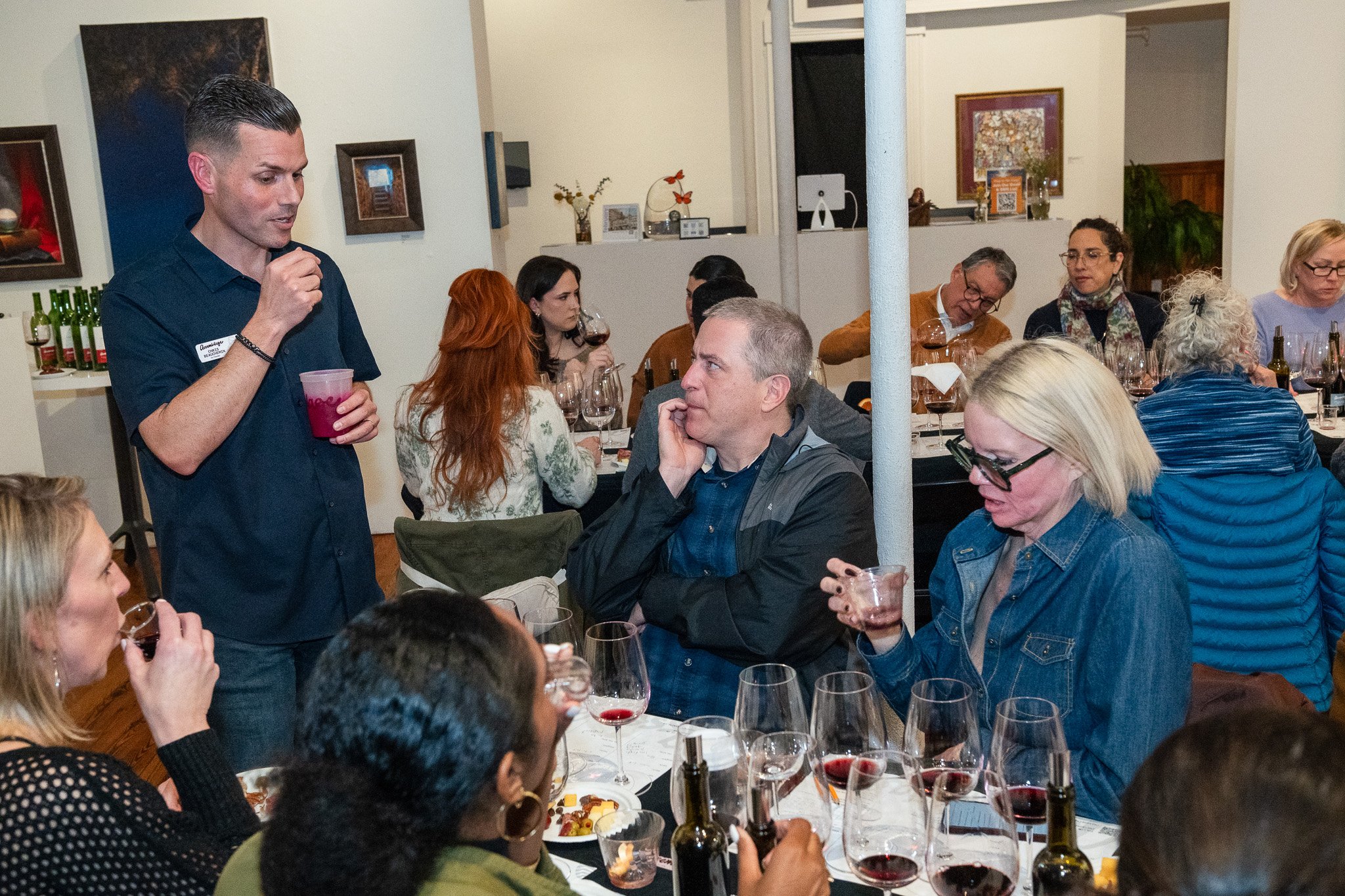 A man giving a wine tasting or speech to a group of people seated around tables with glasses of wine, food, and bottles in a cozy indoor setting.