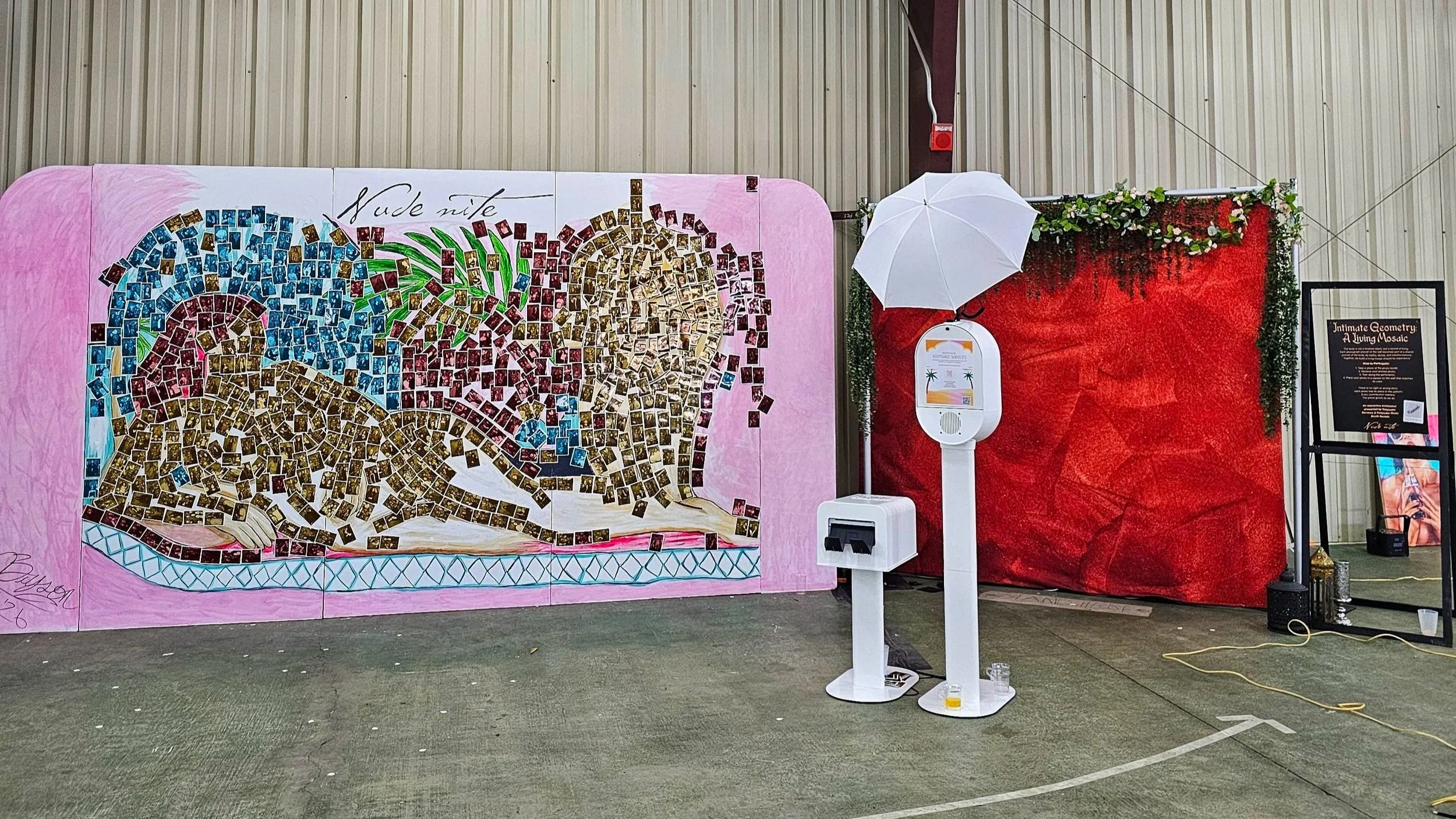 An indoor scene with a colorful mosaic artwork of a reclining nude woman, a white umbrella, a photo booth, a red backdrop with greenery, and a framed poster on a black stand.
