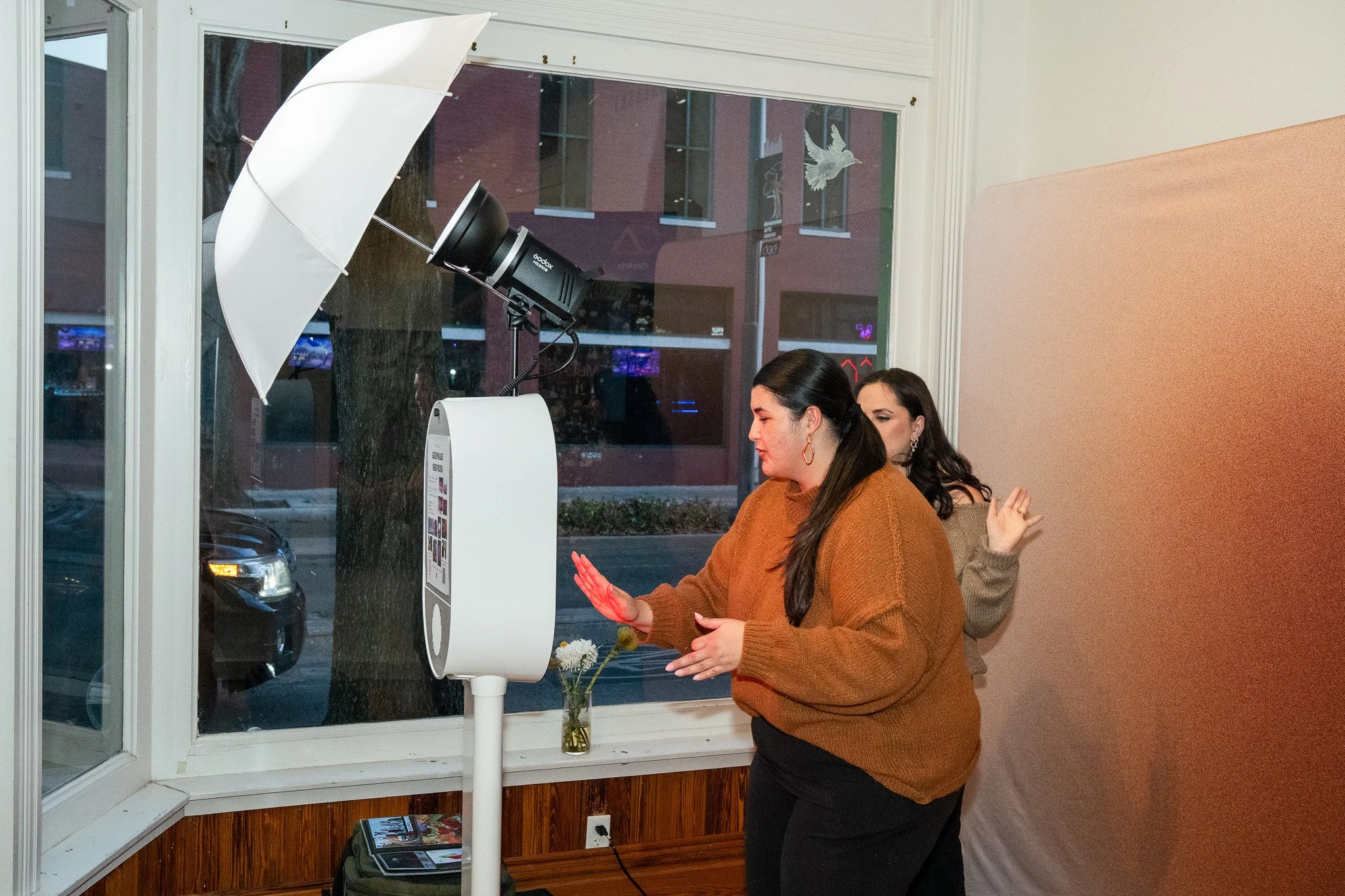Two women are using a photo booth with a camera and a ring light inside a room with a large front window. One woman in a brown sweater is posing with her hand near the photo booth, while the other woman behind her is waving and smiling. There is a sm