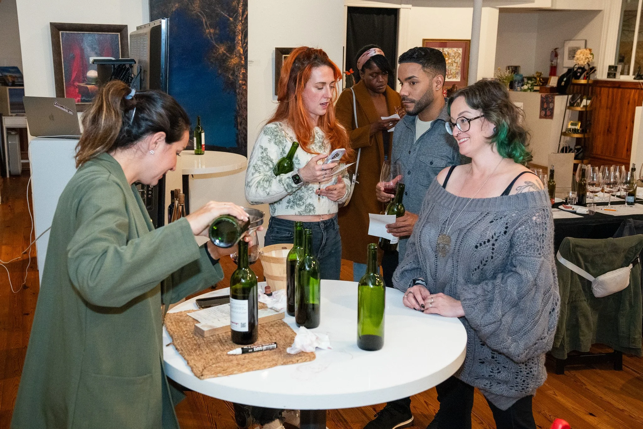 Group of five women gathered around a white table with green wine bottles, glasses, and notes, indoors at a social gathering or wine tasting event.