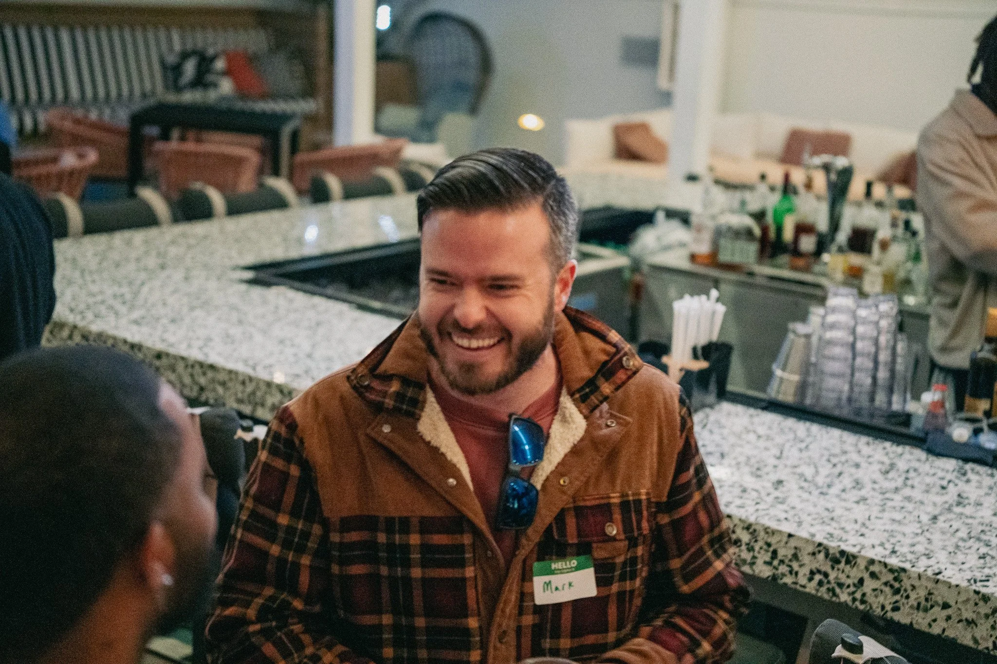 A man with dark hair and a beard, wearing a brown jacket with plaid lining and a pink shirt, smiling and talking to a woman with dark hair tied back, at a bar or cafe counter with drinks and supplies on the countertop.