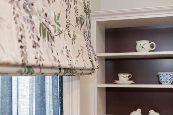 Close-up of a curtain with floral and foliage pattern, partially covering a window, with a white built-in shelf holding cups and bowls in the background.