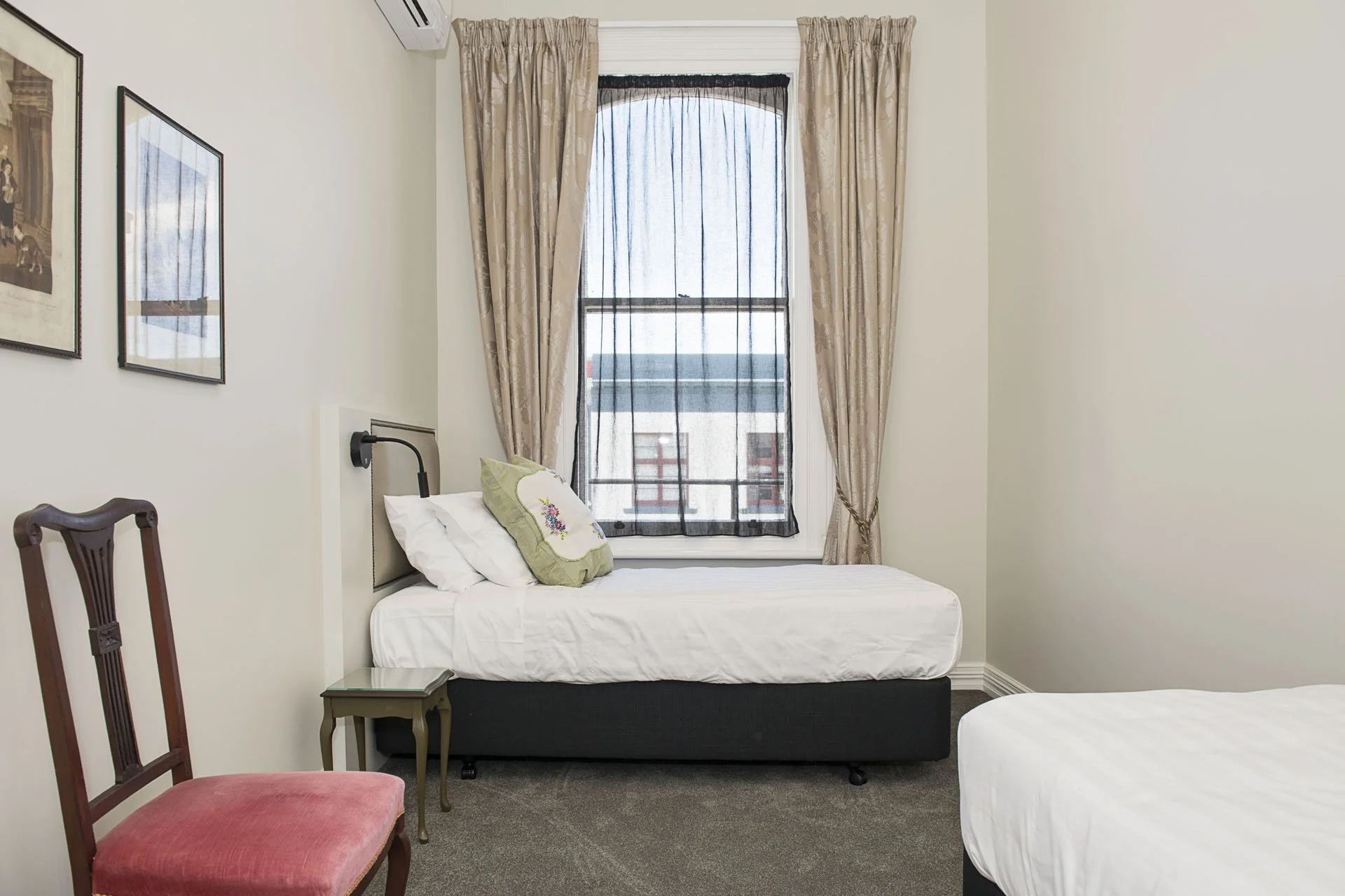 A bedroom with two twin beds, one near a large window with beige curtains and sheer black curtains, black wall-mounted reading lamps, a green embroidered pillow on the bed, a wooden chair with a red upholstered seat, a small gray bedside table, frame