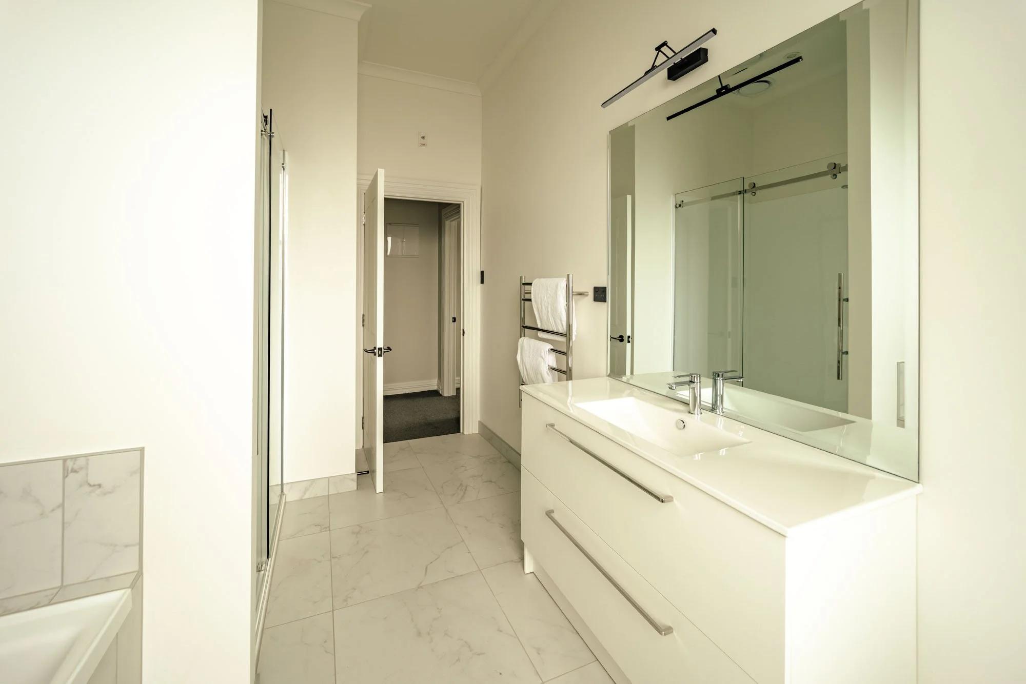 Modern bathroom with white cabinetry, large mirror, shower enclosure, towel rack with towels, and doorway to another room