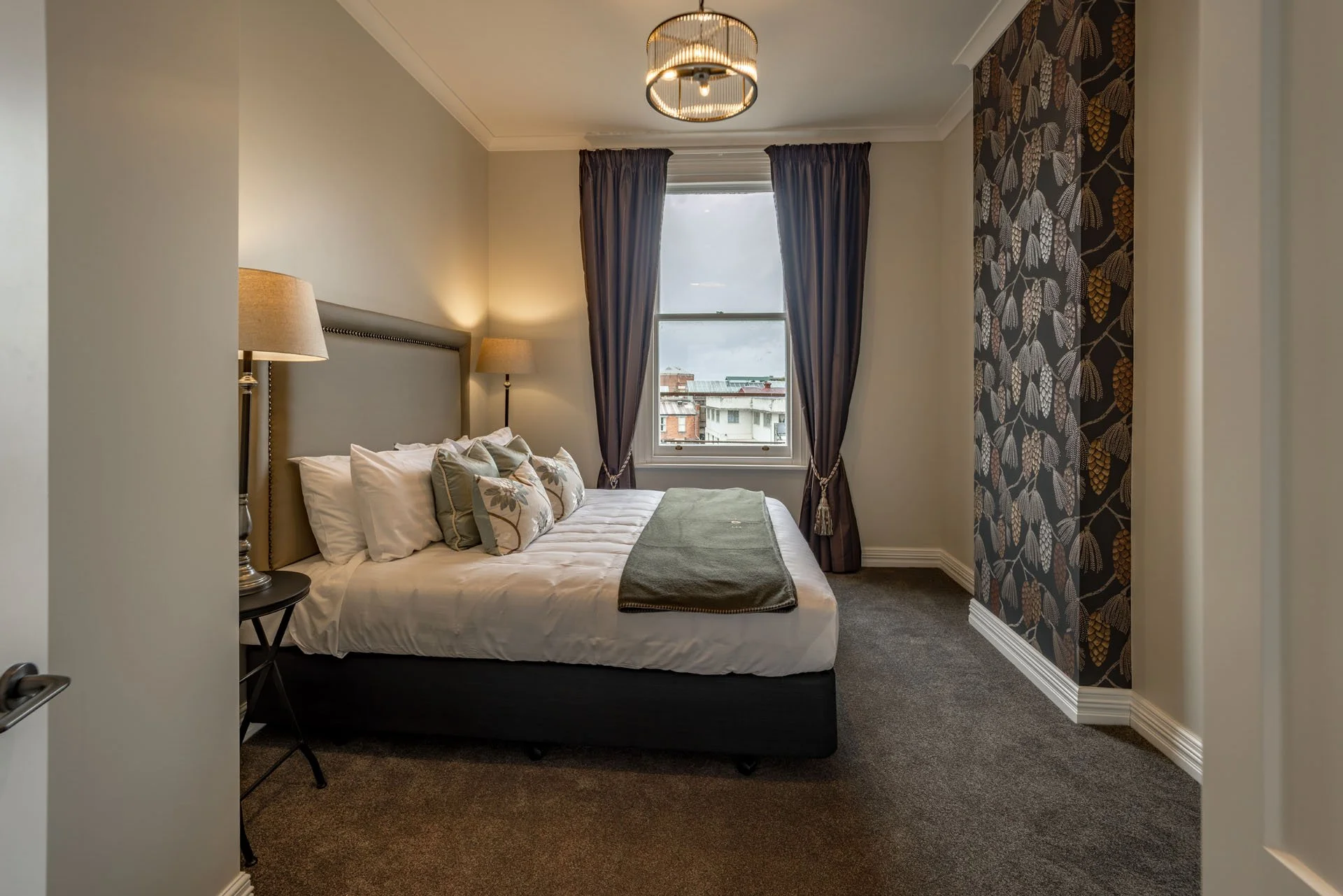 A cozy bedroom with a large bed, nightstands with lamps, a window with dark curtains, and patterned accent wall.