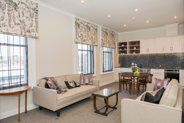 Living room with two beige sofas, decorative pillows, a wooden side table, and a wooden coffee table. Large windows with patterned curtains, and a kitchen area with white cabinets, a gray backsplash, a round dining table, and chairs.