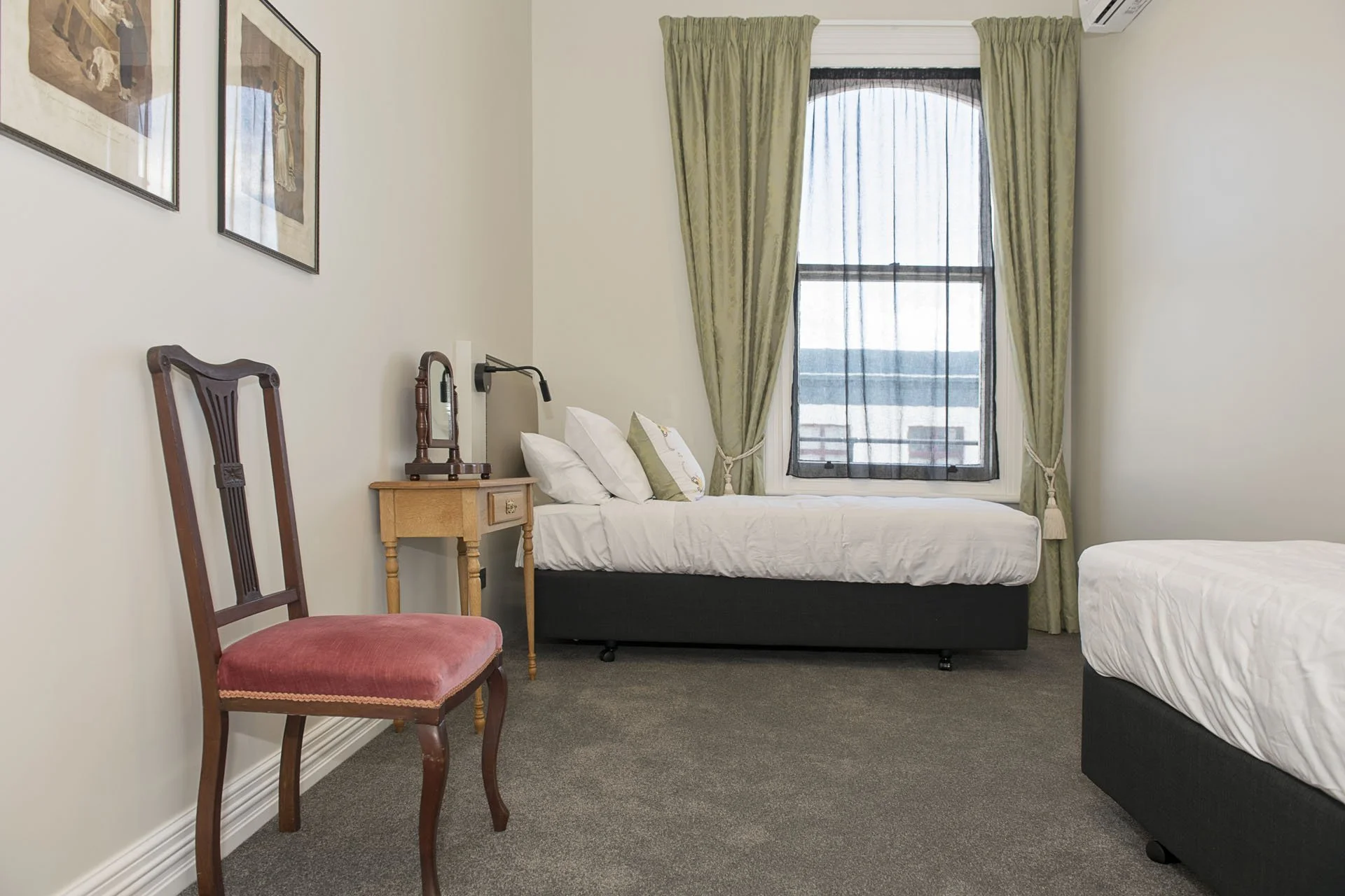 Two beds in a bedroom, one is a single bed with a wooden nightstand, a mirror, and a lamp, and the other is a larger bed, with a window in the background with curtains and sheer layers.