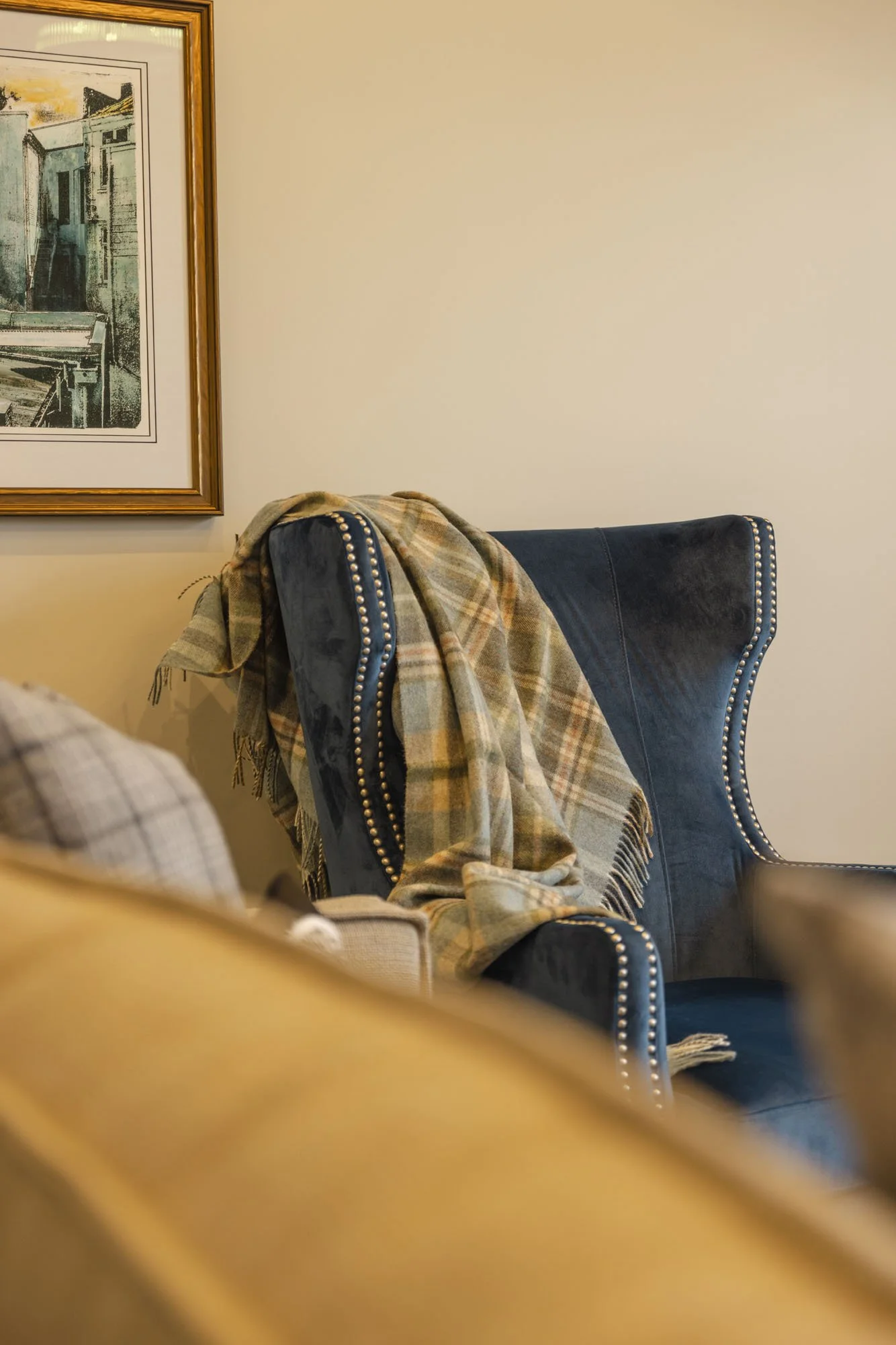 A living room corner with a navy wingback chair that has silver nailhead trim, draped with a plaid throw blanket. A portion of a beige sofa with a plaid fabric is visible in the foreground. A framed painting of an abstract cityscape hangs on the wall nearby.