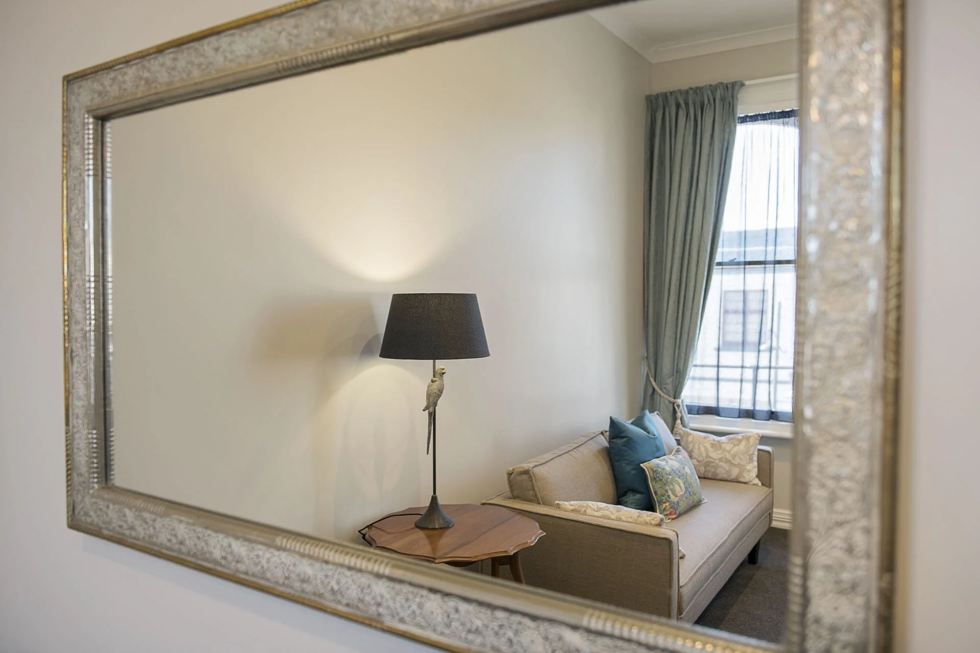 Reflection of a room with a beige sofa, colorful pillows, a window with teal curtains, a side table, and a black lampshade in a large decorative mirror.