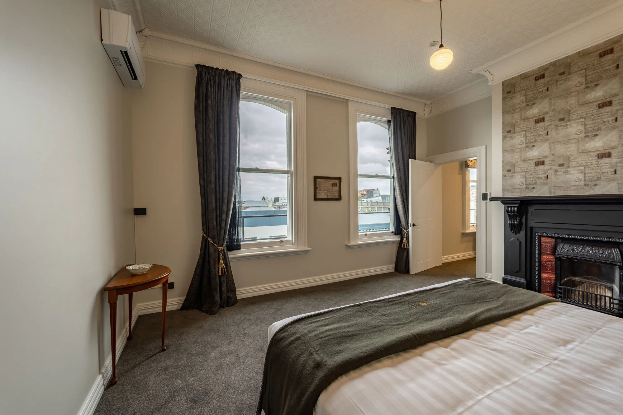 A bedroom with two large windows, dark curtains, a bed with a white and beige striped duvet, a black fireplace surround, a small wooden side table with a bowl, and a ceiling light fixture.