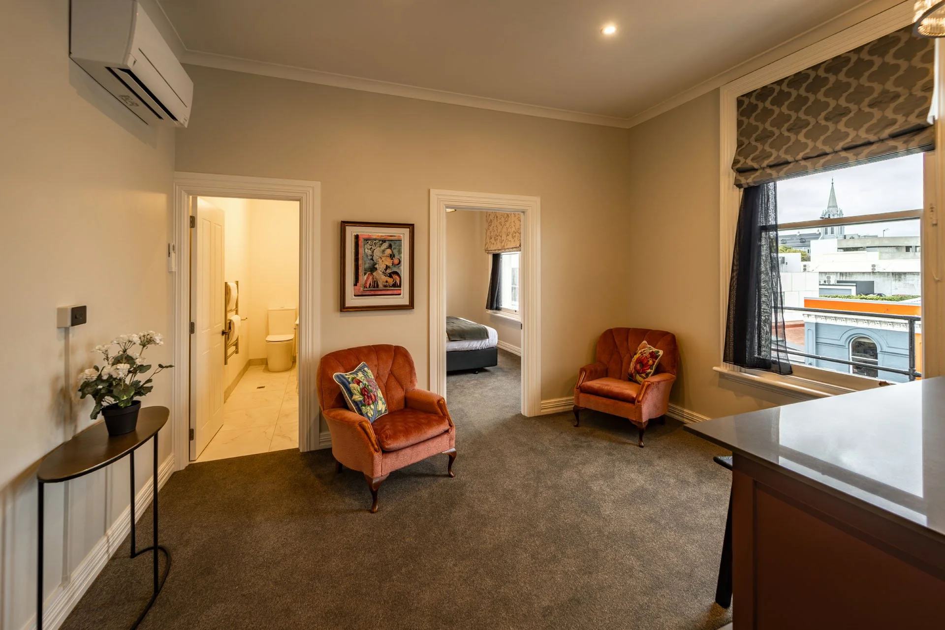Interior of a hotel room with a large window, two upholstered armchairs, a small table with a potted plant, a picture on the wall, and a view of a cityscape with a church steeple outside.