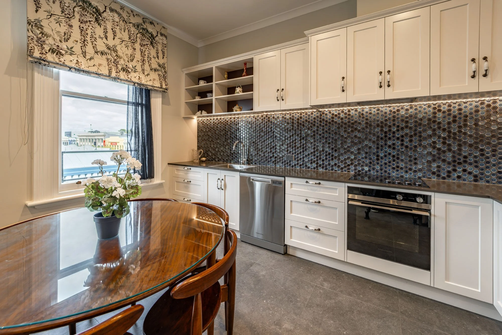 Kitchen with white cabinets, a stainless steel dishwasher, built-in oven, and copper hexagonal tile backsplash. A wooden dining table with chairs and a potted plant by the window.