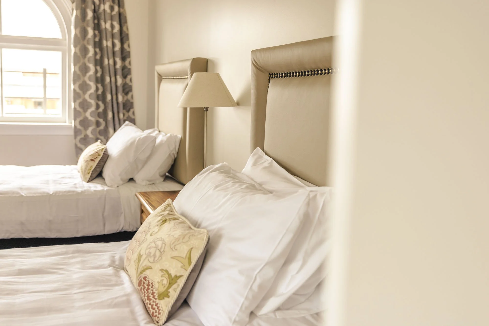 A hotel room with two beds, beige headboards, white bedding, and decorative pillows, with a window in the background.