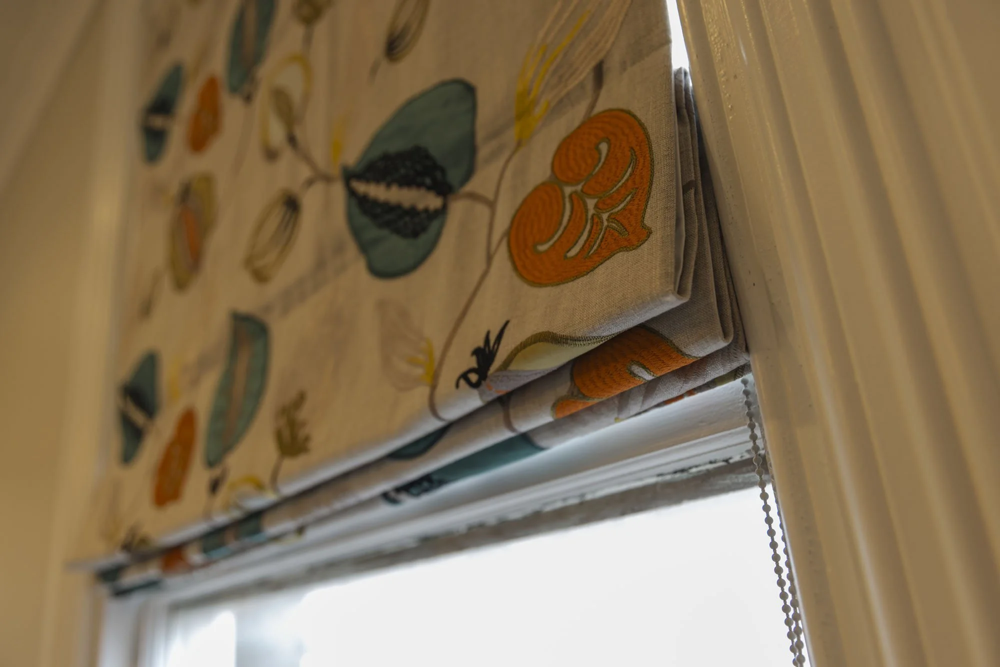 Close-up of a fabric window shade with colorful embroidered leaves and flowers, partially rolled up, next to a white window frame.