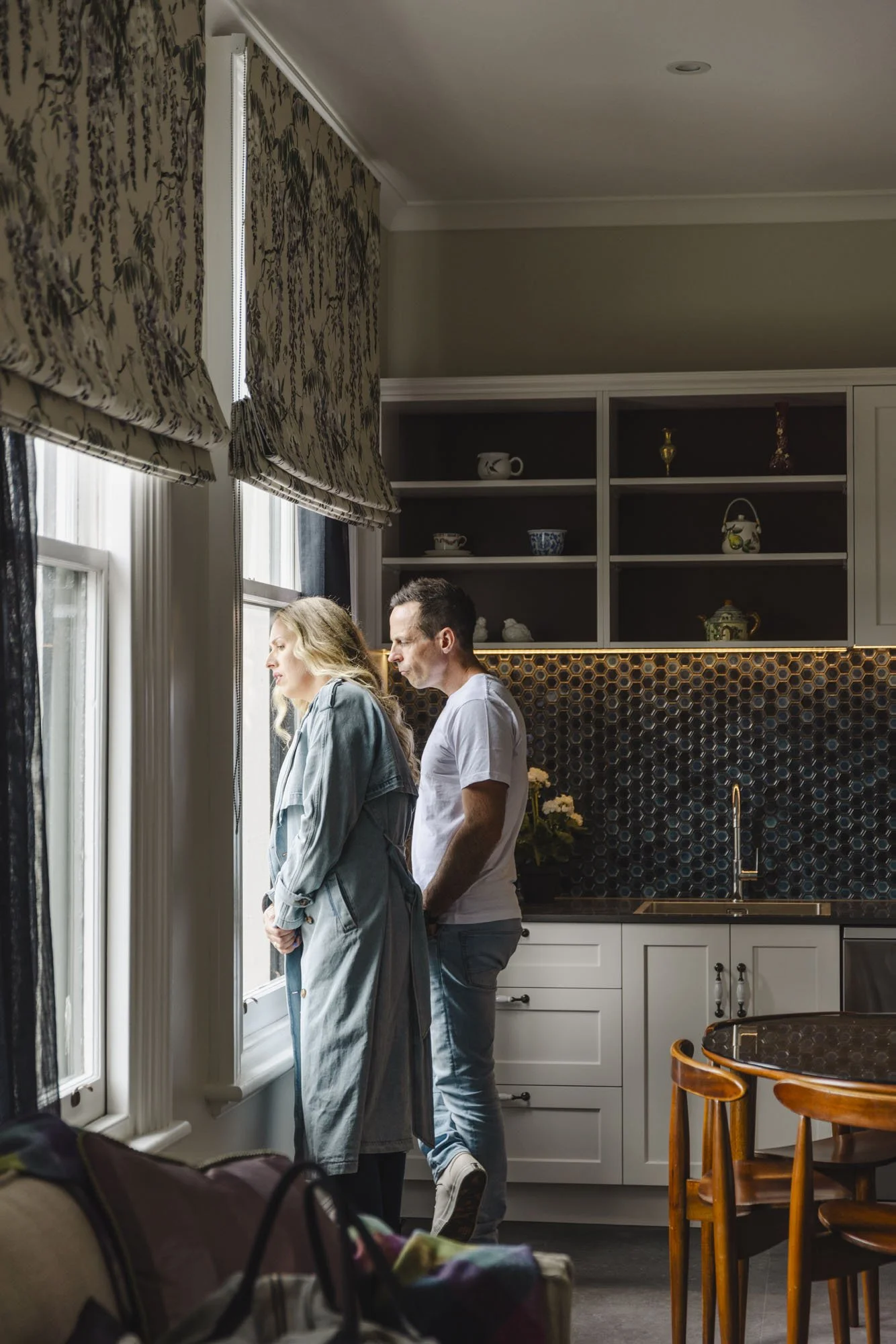 A man and a woman standing near a window in a kitchen, looking outside with contemplative expressions.