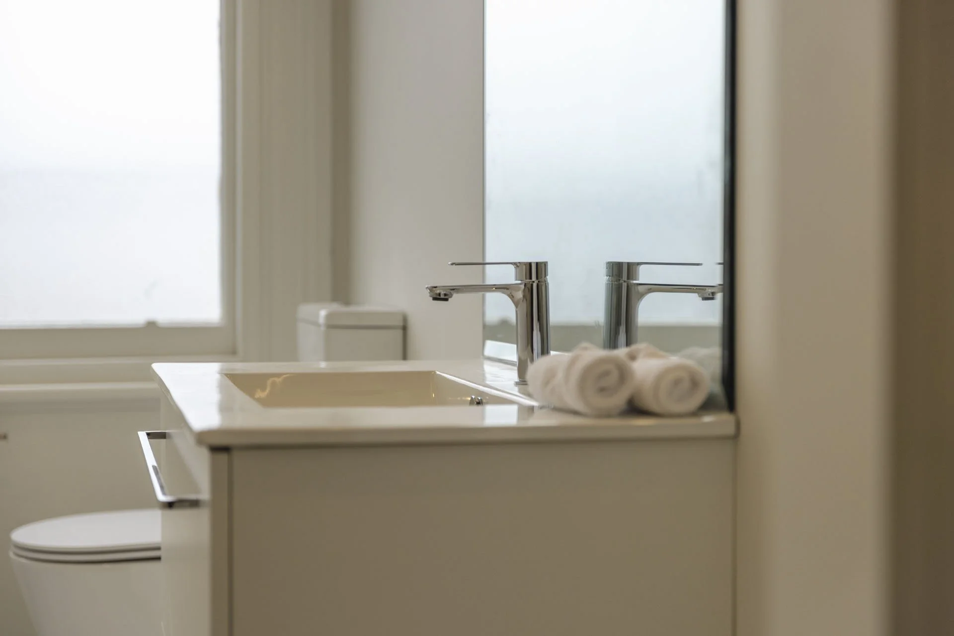 Bathroom sink with rolled towels and a reflection in the mirror, with a window in the background.