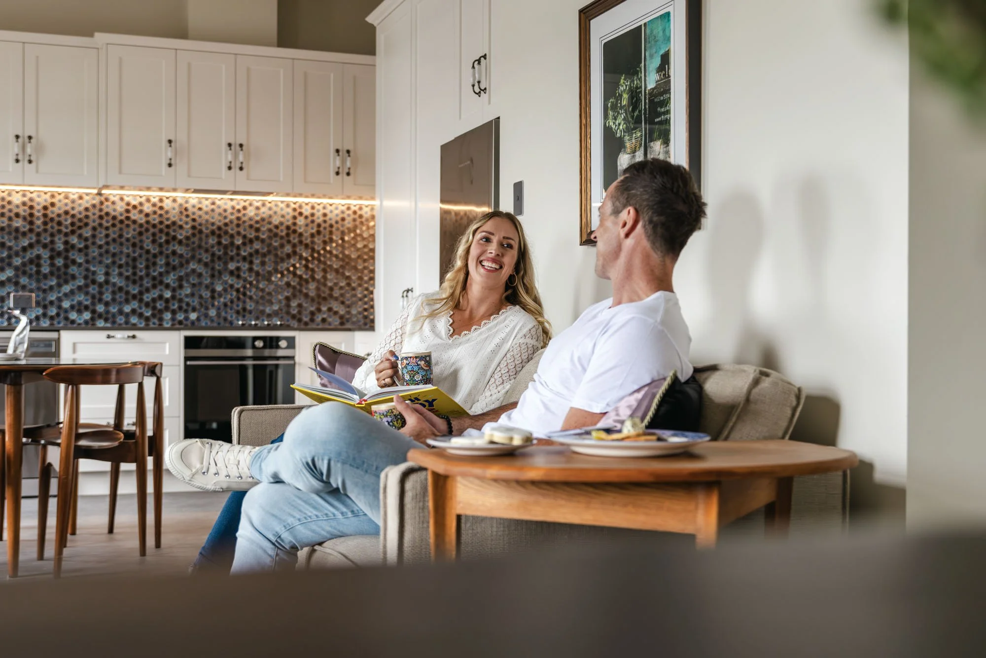 A man and woman sitting on a sofa in a cozy kitchen, smiling and talking, with the woman holding a coffee mug and the man holding a book.