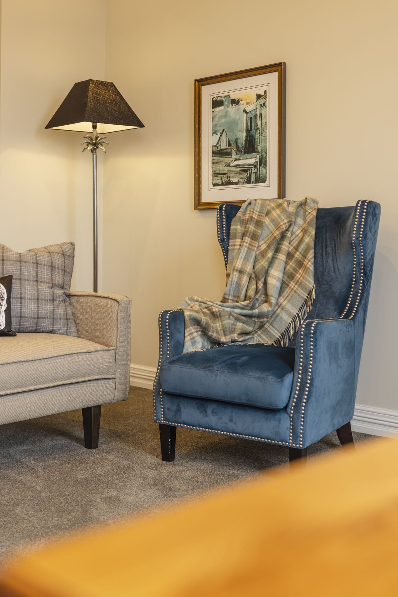 A living room corner featuring a beige sofa with cushions, a blue velvet wingback chair with nailhead trim draped with a plaid blanket, a black floor lamp, and framed artwork on the wall.