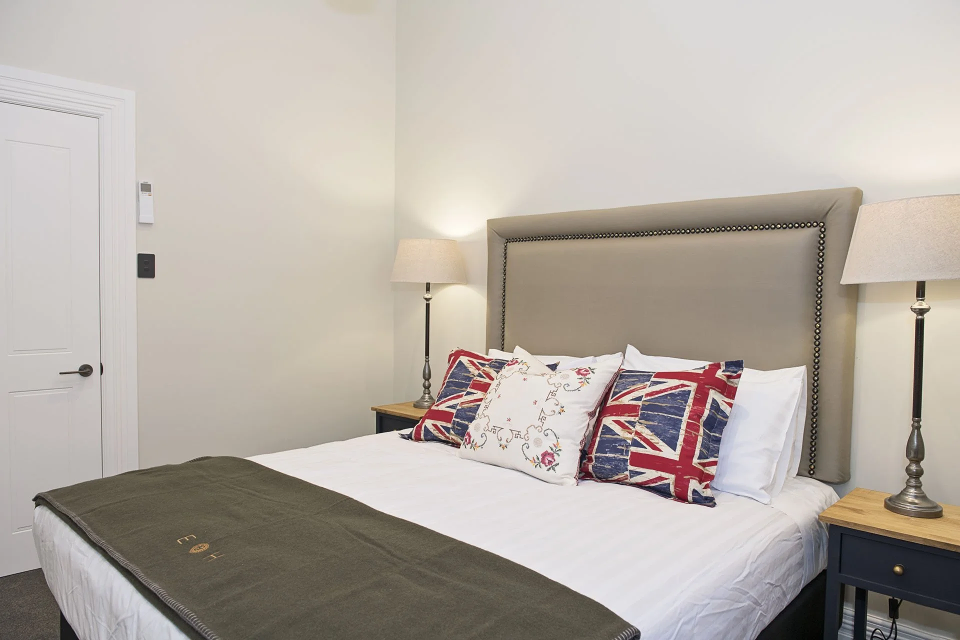 A neatly made bed with a beige upholstered headboard, multiple pillows with Union Jack and floral designs, white sheets, a brown bed throw, and two matching bedside lamps in a cozy bedroom.