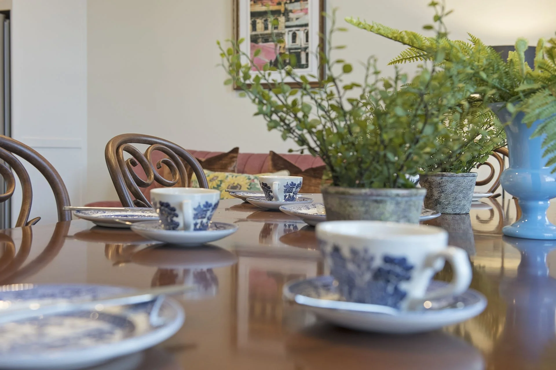 Dining table set with blue and white patterned cups and plates, and a large plant in a gray pot, with a sofa and framed picture in the background.