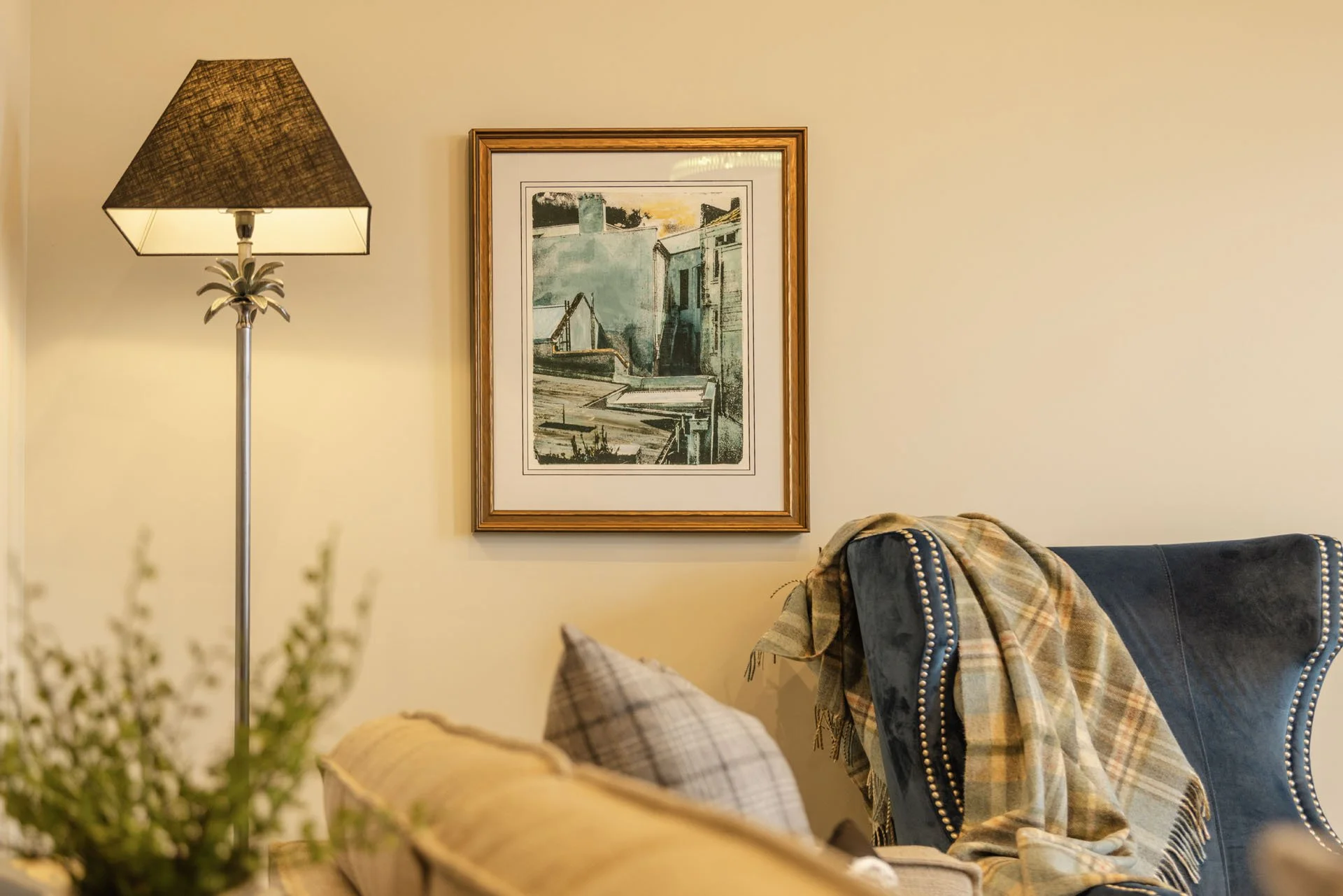 Living room wall with a framed artwork, a tall standing lamp with a pineapple-inspired base, a plaid blanket draped over a blue armchair, and a beige sofa with checkered cushions.