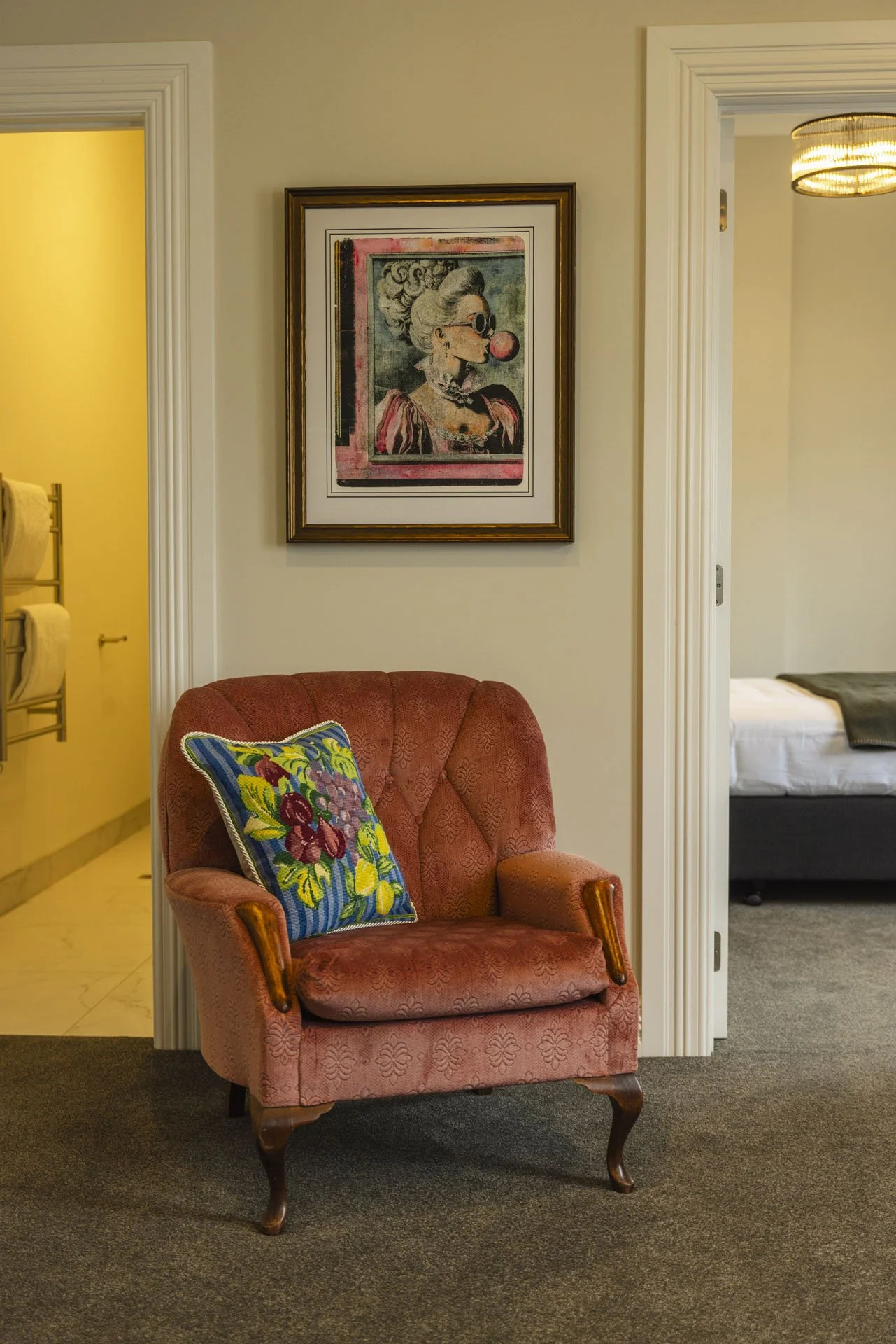 A cozy room corner with a vintage red armchair, floral pillow, framed modern artwork of a woman with a bubblegum, and view into a bedroom and bathroom.